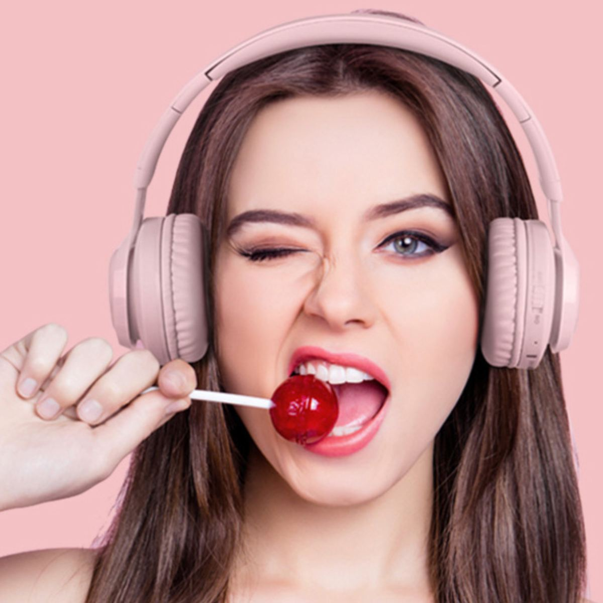 Une femme aux longs cheveux bruns porte un casque rose tout en faisant un clin d'œil et en tenant une sucette dans sa bouche.