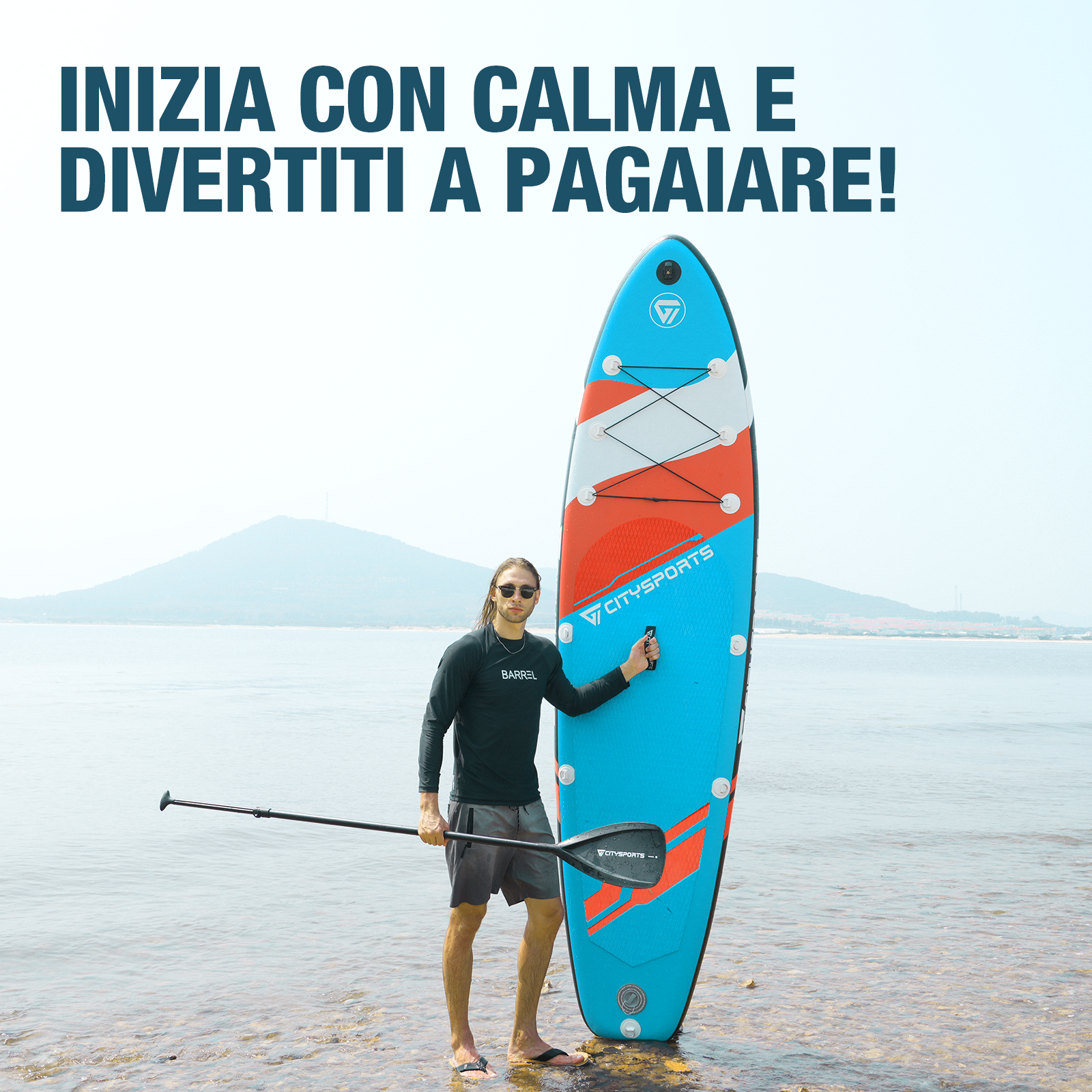 Un uomo è in piedi con una tavola da paddle nelle acque poco profonde di uno specchio d'acqua, con una montagna sullo sfondo.