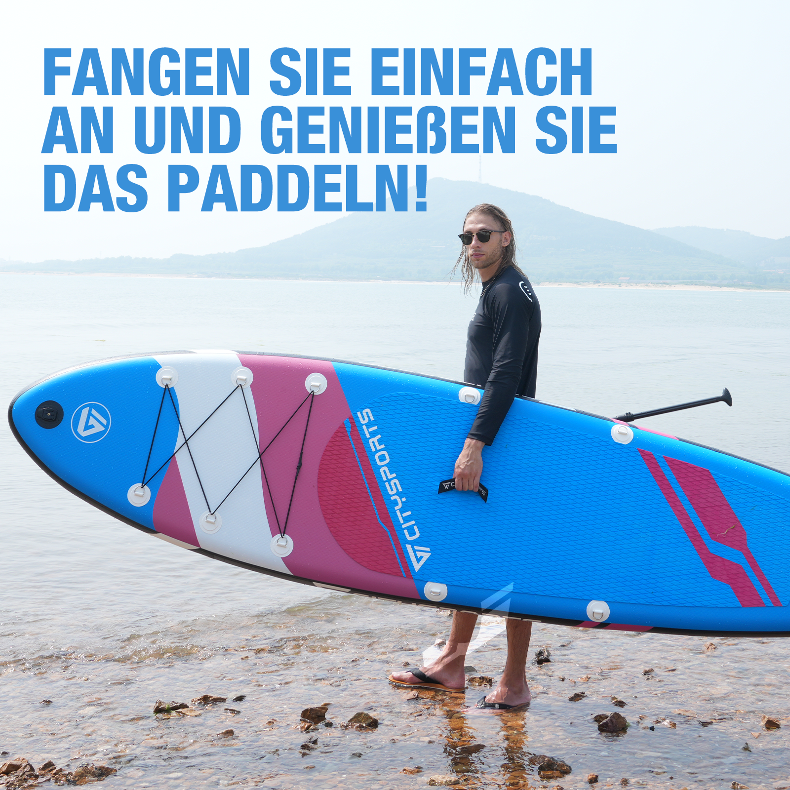 Ein Mann trägt ein blau-pinkes Paddleboard an einem felsigen Strand neben Wasser mit Bergen im Hintergrund.