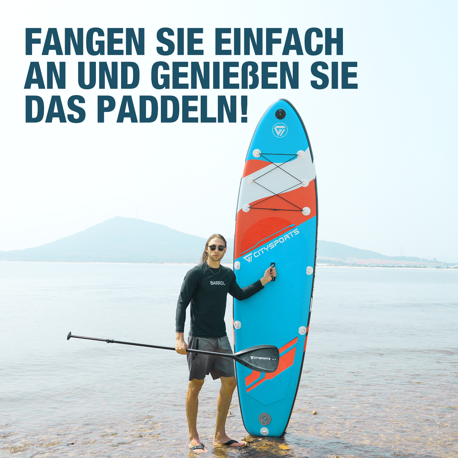 Ein Mann steht an einem Strand und hält ein blaues und orangefarbenes Paddleboard, in der Nähe von Wasser und Bergen.