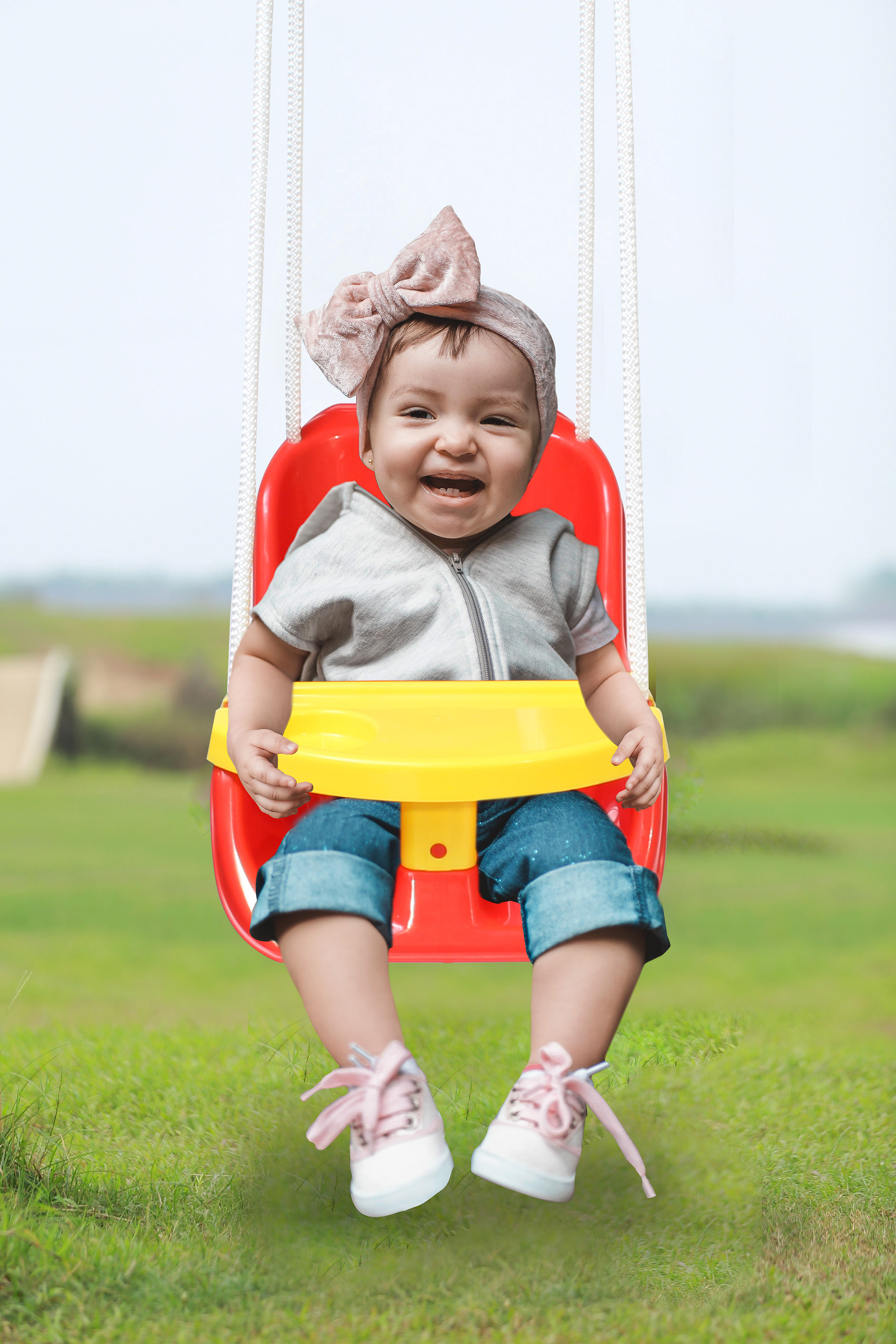 Ein Baby in einer rot-gelben Schaukel, lächelnd, mit Schleife auf dem Kopf, auf grünem Grund.