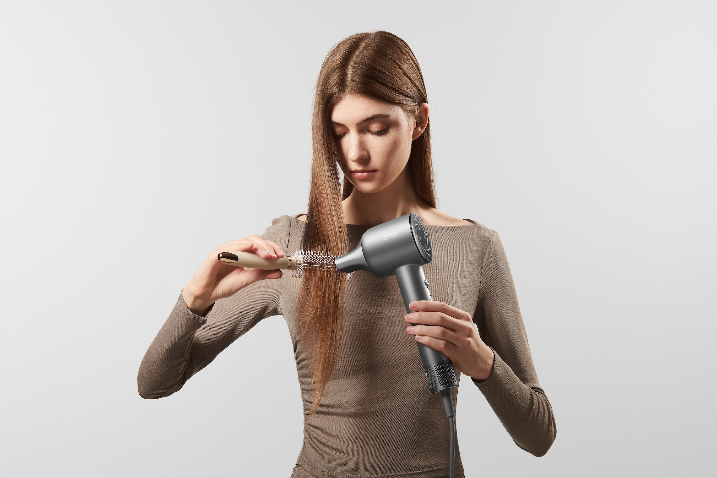 Eine Frau bürstet ihre langen braunen Haare mit einer Haarbürste, während sie einen grauen Föhn hält, vor weißem Hintergrund.