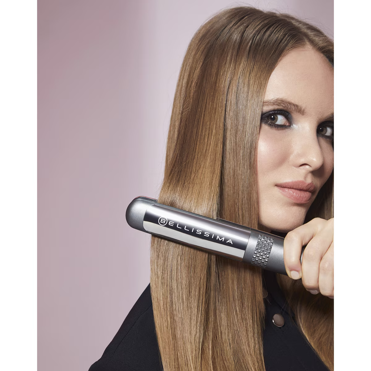 Kobieta używająca prostownicy na długich, jasnobrązowych włosach, patrząca w kamerę.