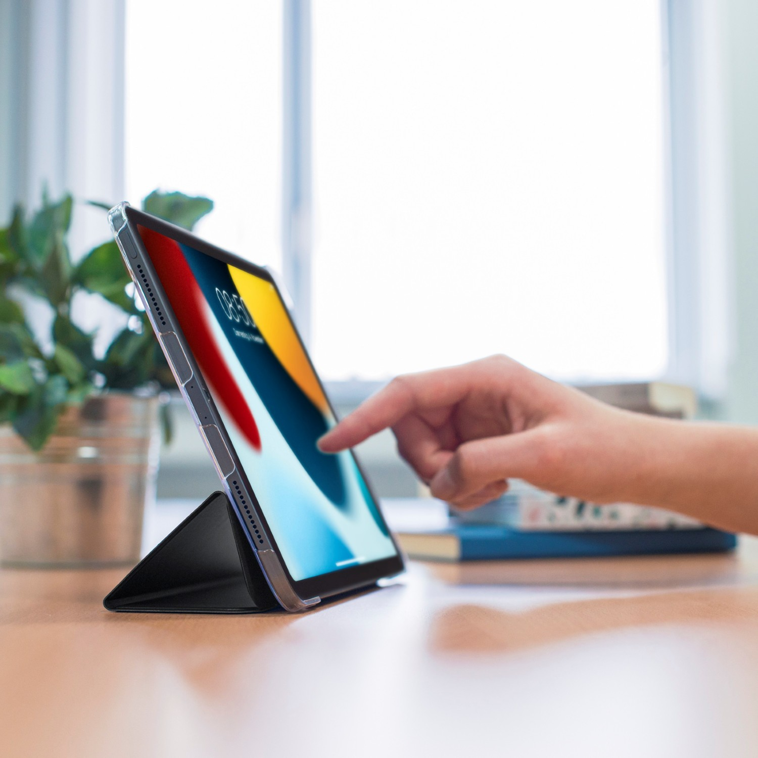 Dłoń dotyka ekranu tabletu na biurku. W tle okno.