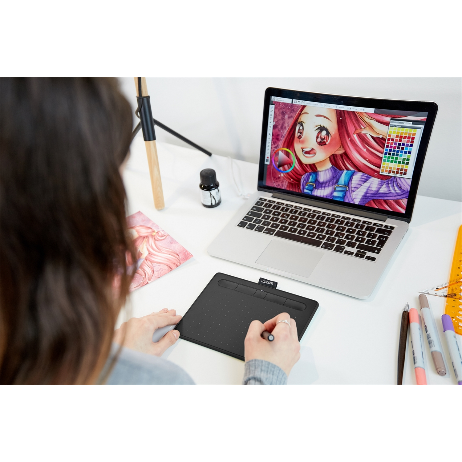 Una persona disegna su un tablet collegato a un laptop. Penne, pennarelli e inchiostro sulla scrivania.