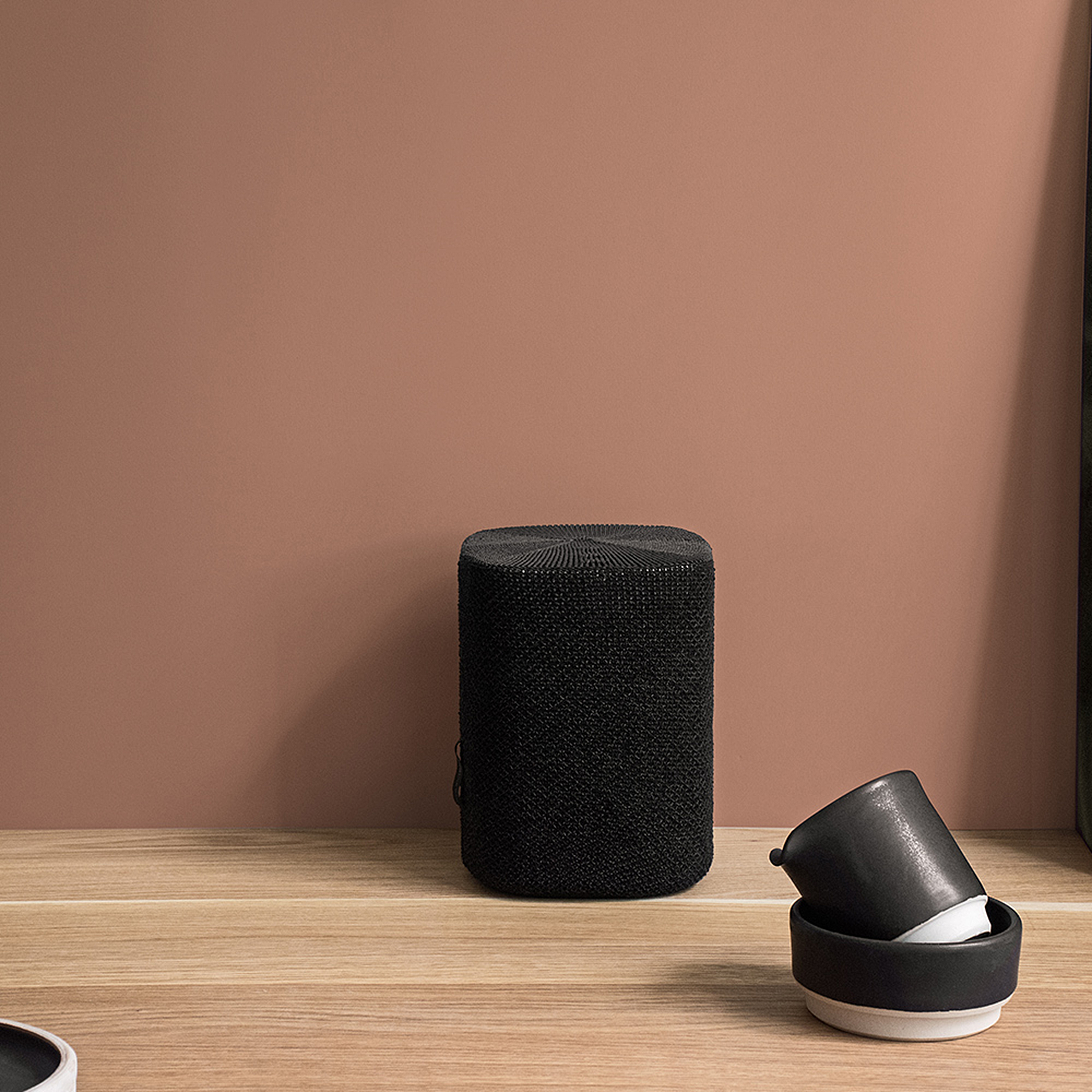 Un haut-parleur noir sur une surface en bois, contre un mur marron. Deux petites tasses noires sont à proximité.