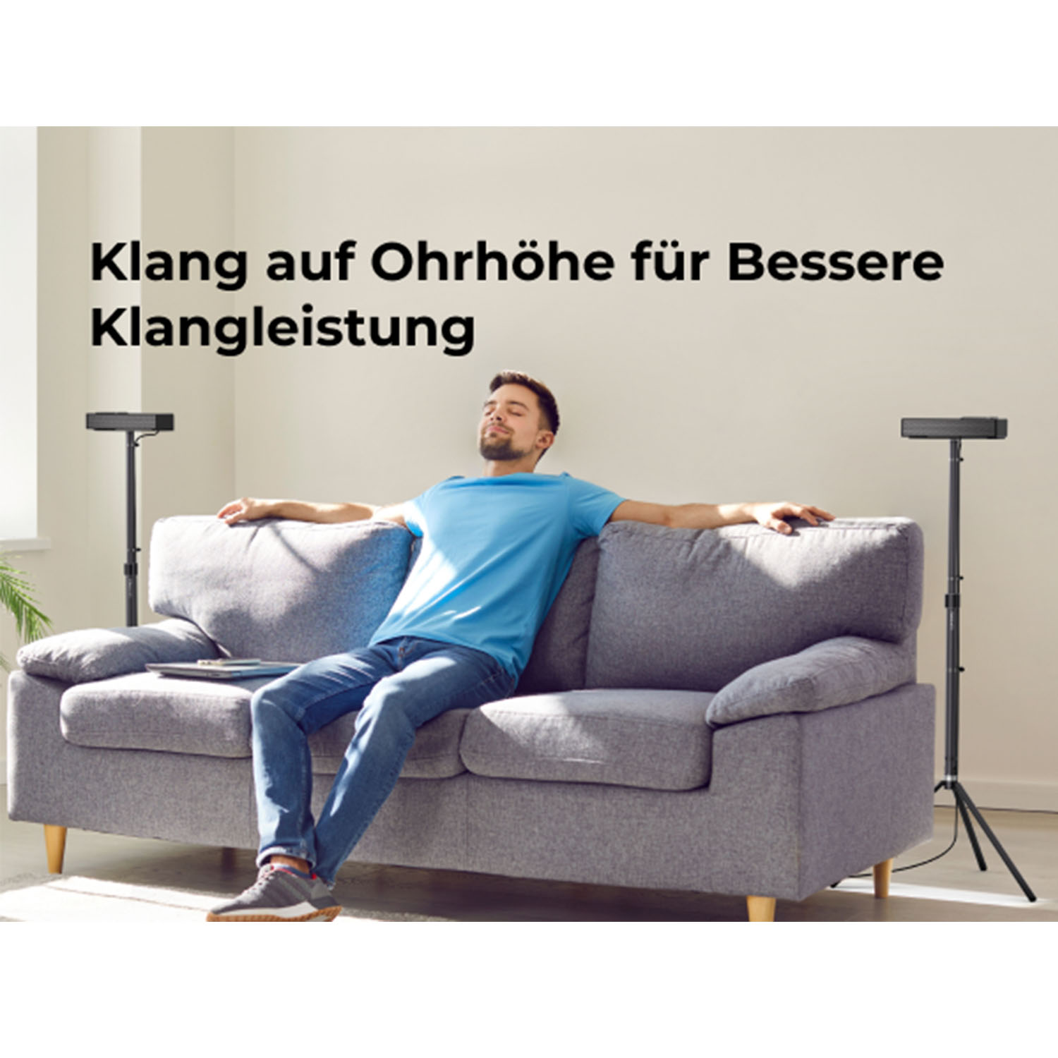 Ein Mann liegt mit ausgestreckten Armen auf einem grauen Sofa. Zwei Lautsprecherständer stehen daneben.