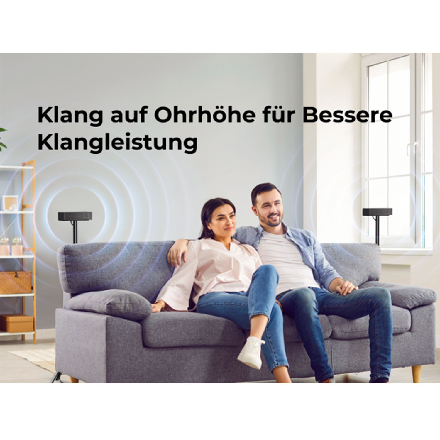 Ein Paar sitzt auf einem grauen Sofa mit zwei Lautsprecherständern. Die Ständer haben Lautsprecher.