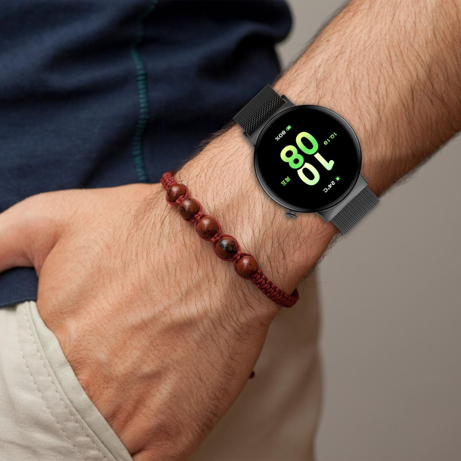 Das Handgelenk einer Person mit einer schwarzen Smartwatch, die Zeit und Akkulaufzeit anzeigt. Ein rotes Perlenarmband wird getragen.