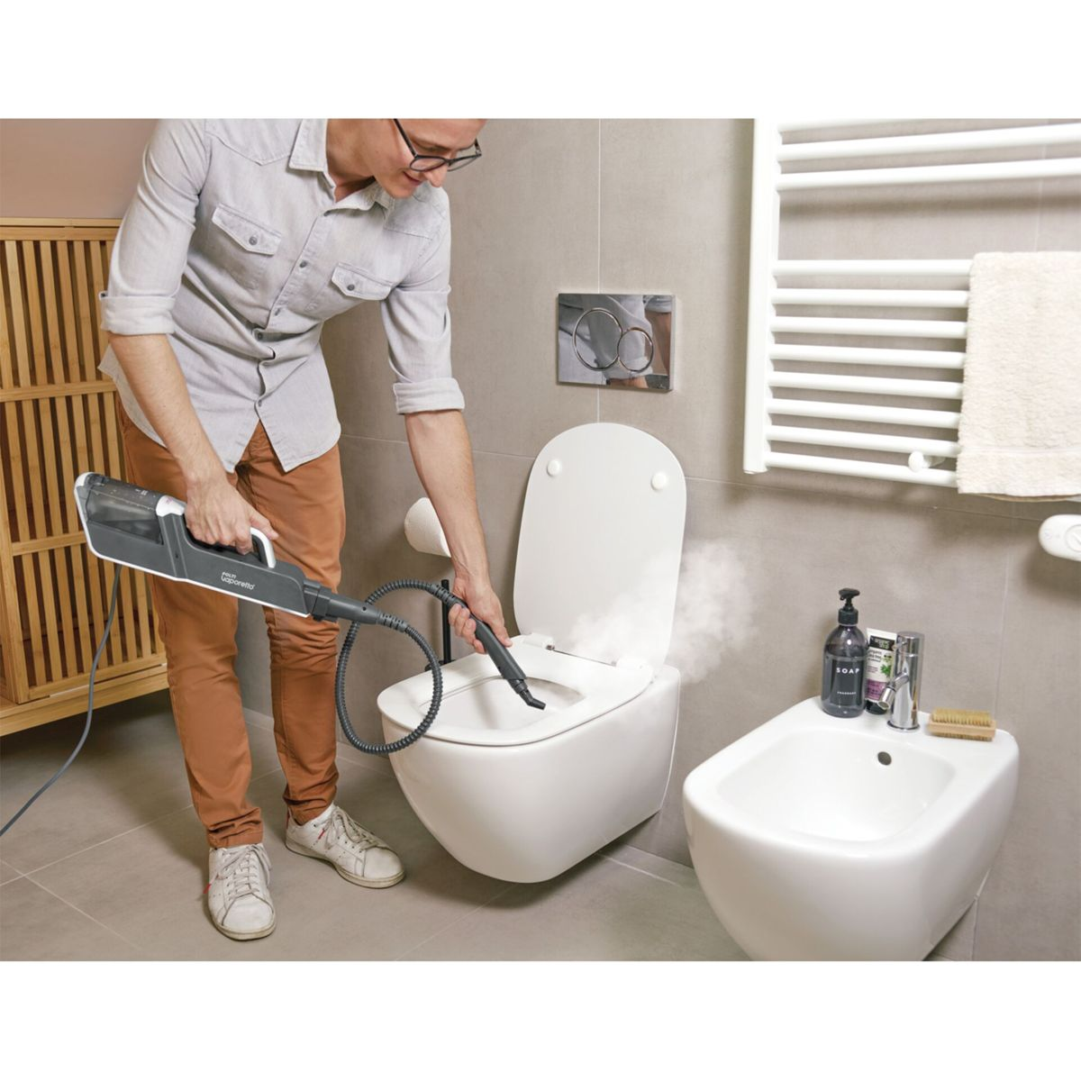 Mężczyzna czyści toaletę parownicą. Deska sedesowa otwarta, obok bidet.