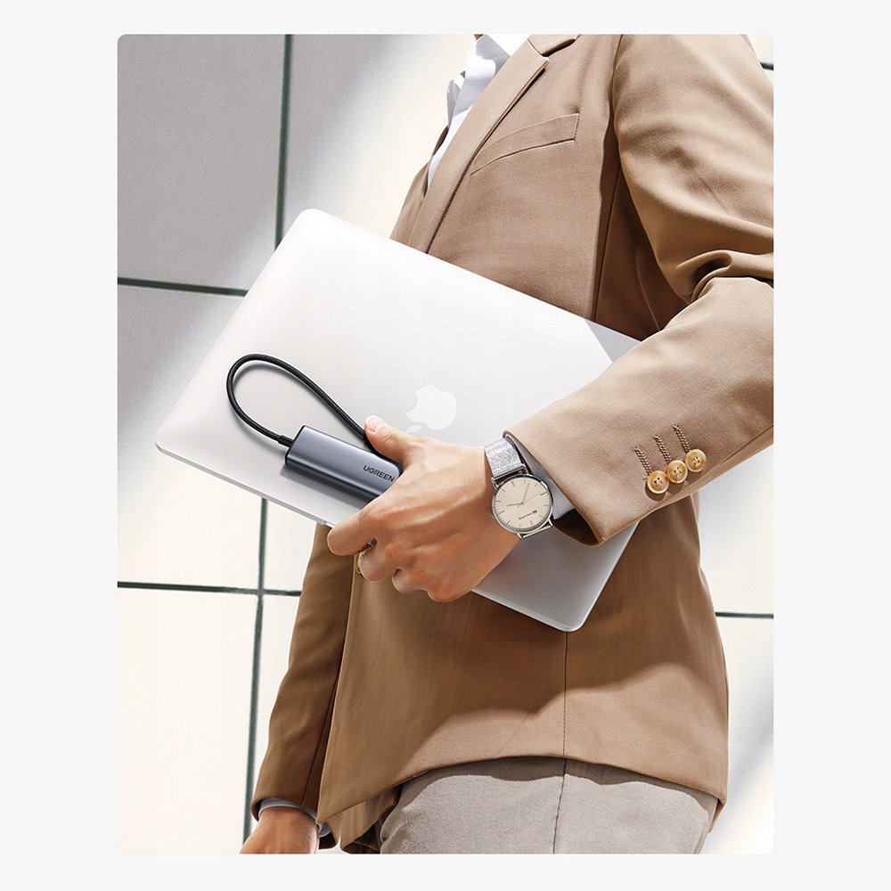 Osoba trzymająca laptopa i hub USB, mająca na sobie zegarek, z jasnym tłem.