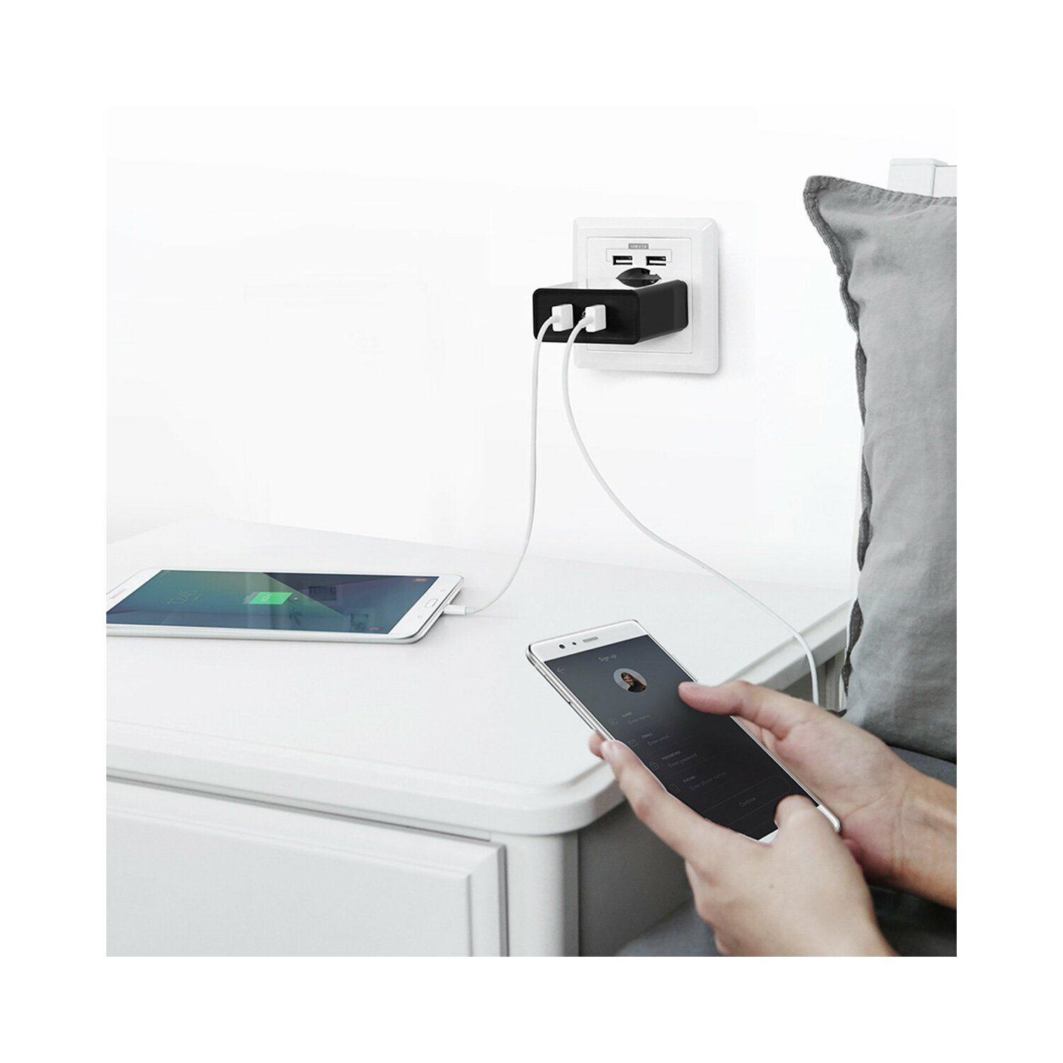 Białe gniazdo ścienne z wpiętą czarną ładowarką, ładujące tablet i telefon na białej szafce nocnej.
