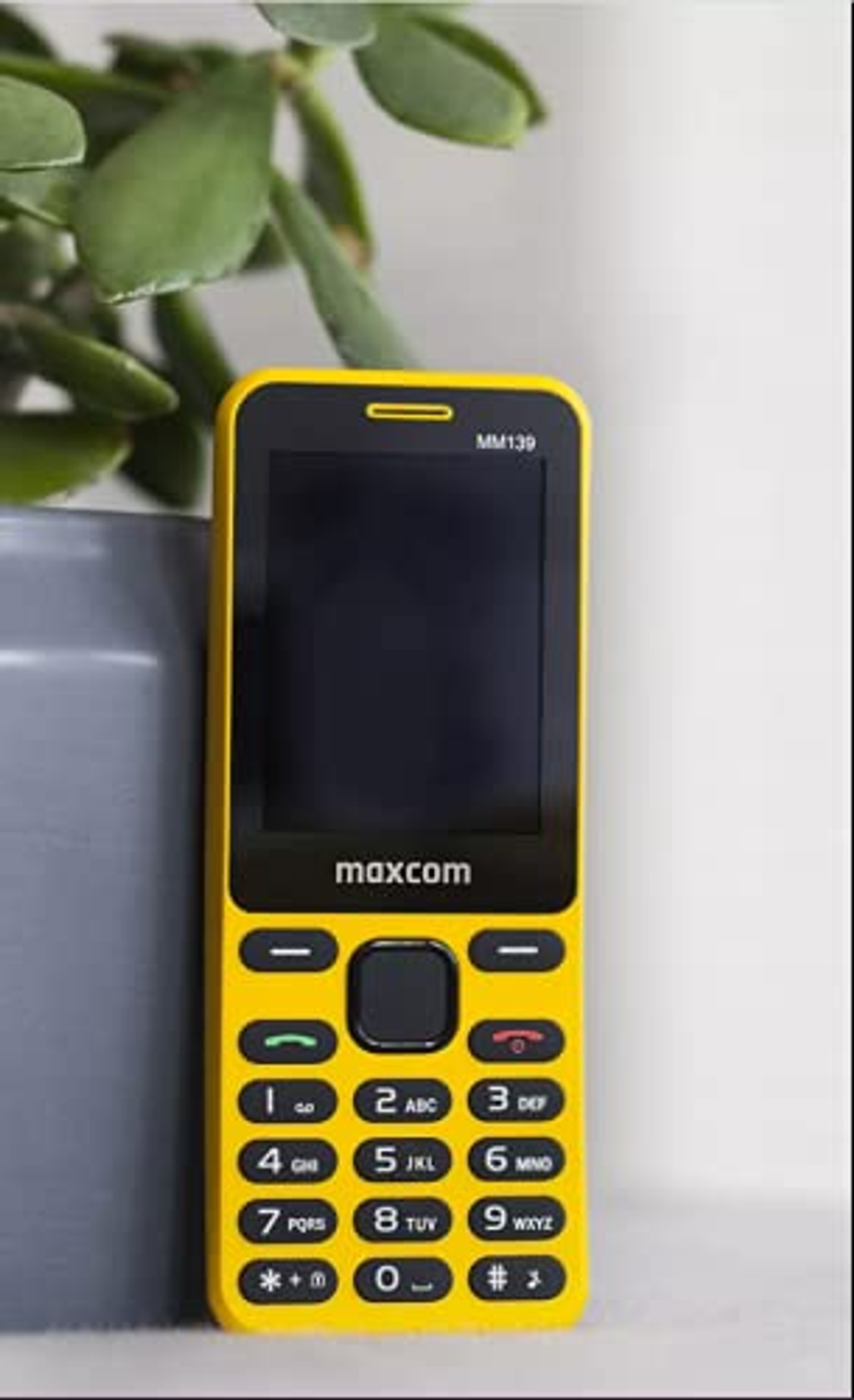 Teléfono Maxcom amarillo, pantalla y teclado negros. Muestra texto MM139. Planta verde al fondo.
