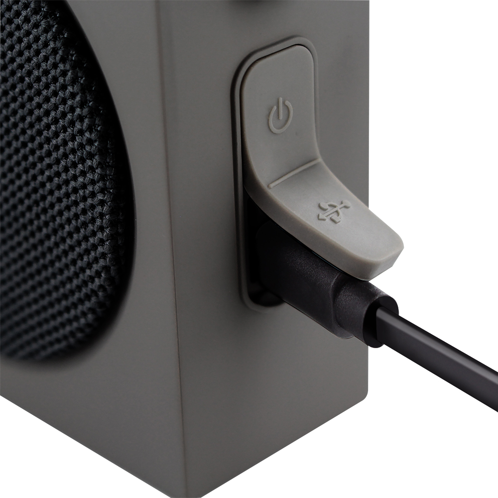 Primer plano de un altavoz gris con un cable de carga negro enchufado. Un botón de encendido es visible.