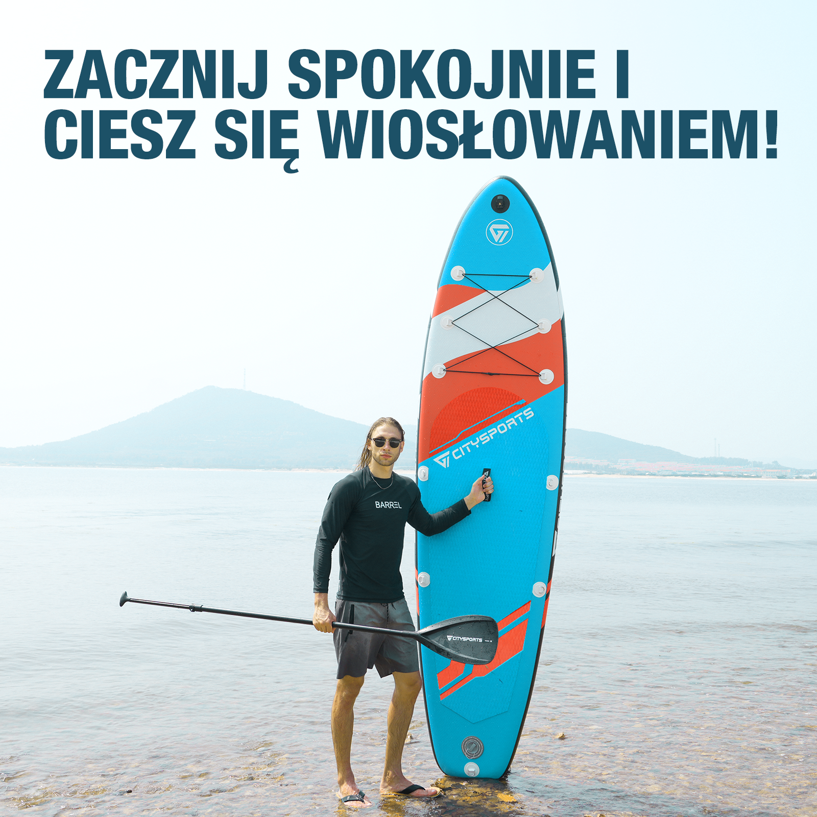 Mężczyzna w okularach przeciwsłonecznych stojący na plaży z deską do wiosłowania, trzymający wiosło. Góra w tle.