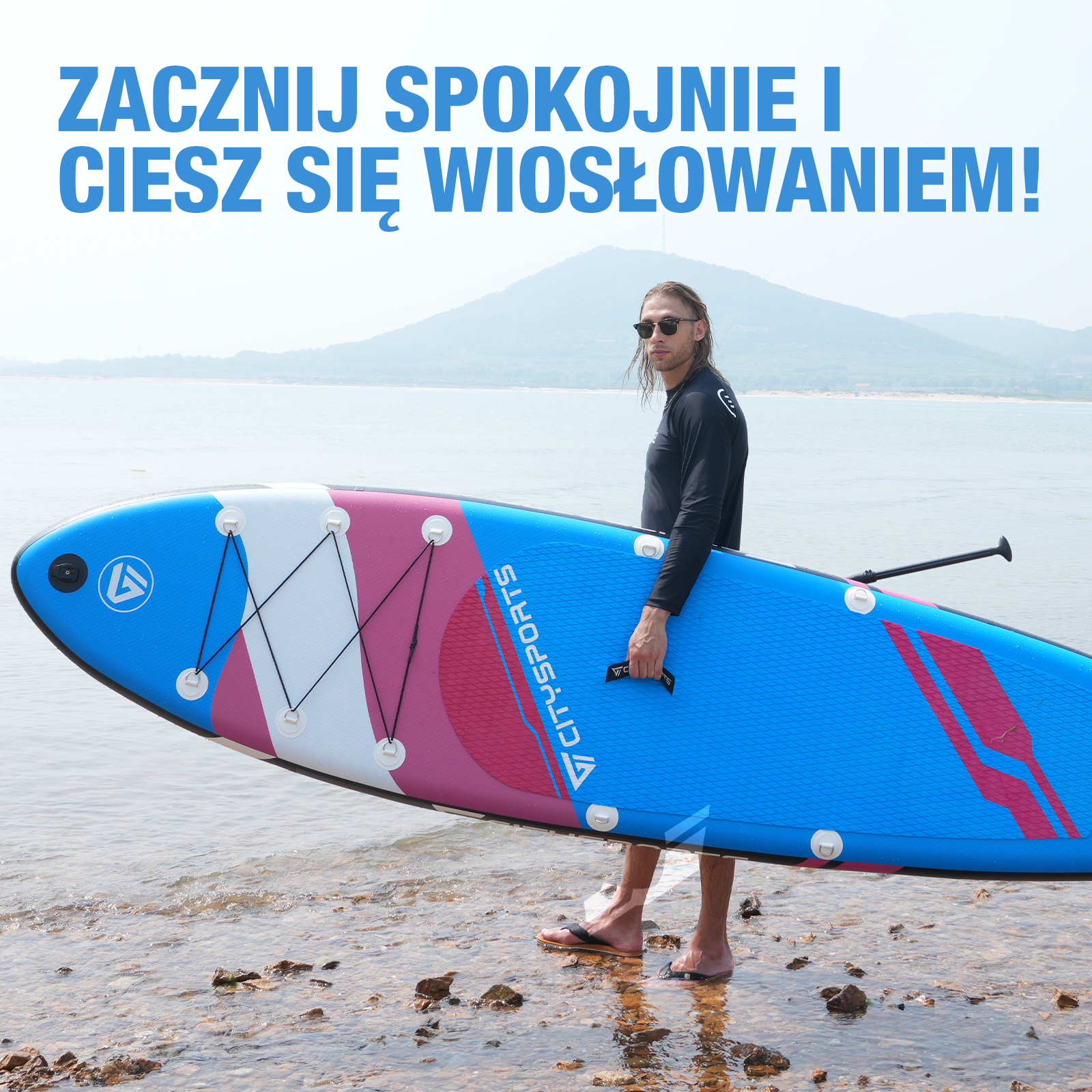 Mężczyzna trzymający deskę do wiosłowania na plaży, z górą i morzem w tle.