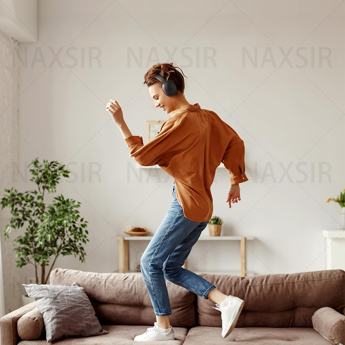 Frau tanzt auf einem Sofa, trägt Kopfhörer, ein orangefarbenes Hemd und Jeans in einem Wohnzimmer.