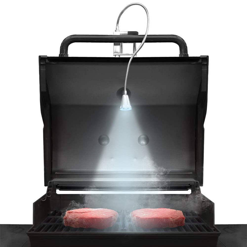 Grill mit zwei Steaks, beleuchtet von einer verstellbaren Lampe, Rauch steigt auf. Weißer Hintergrund.