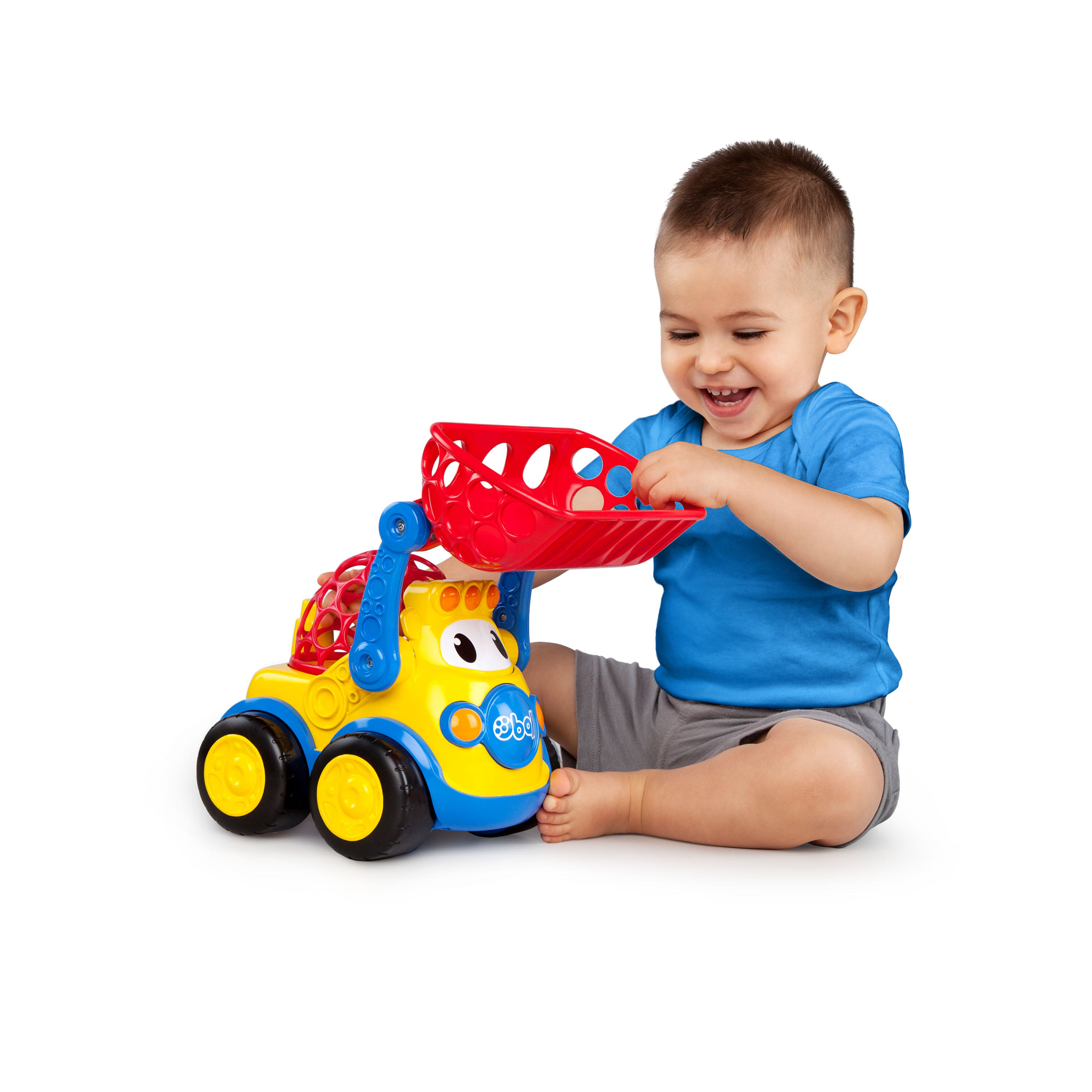 Un niño pequeño juega con un camión de juguete amarillo, azul y rojo. Sonríe, sentado en blanco.