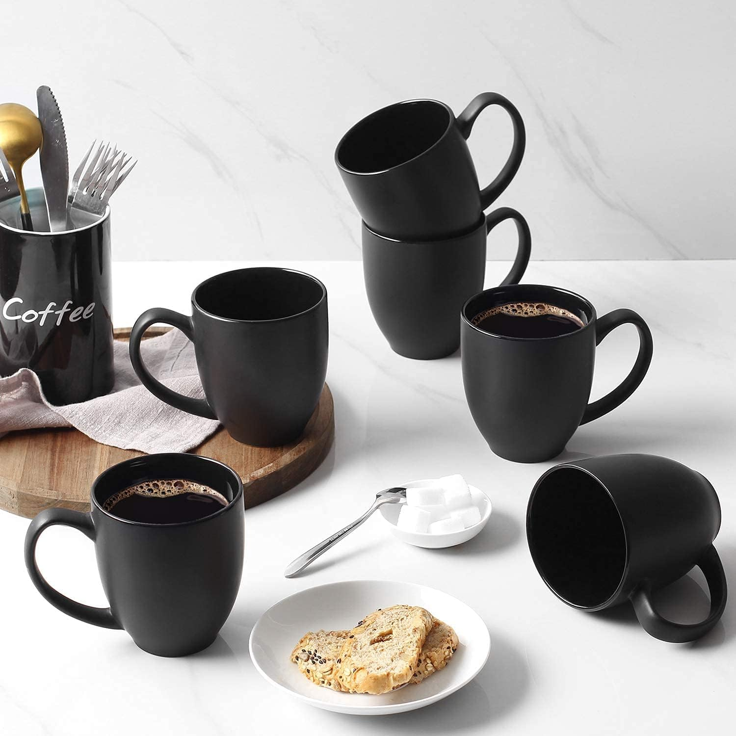 Diverse tazze nere con caffè su una superficie di marmo. Sono presenti un cucchiaio e zollette di zucchero.