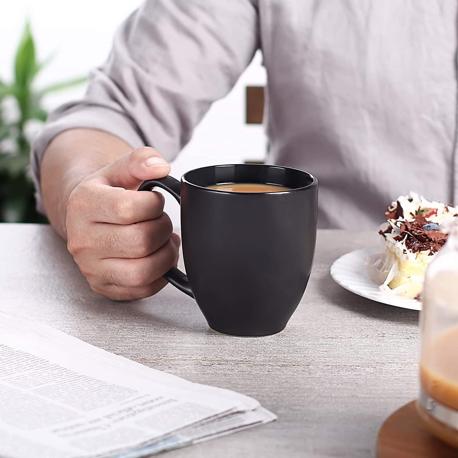 Una persona tiene in mano una tazza nera con caffè. Un giornale e una fetta di torta sono vicini.
