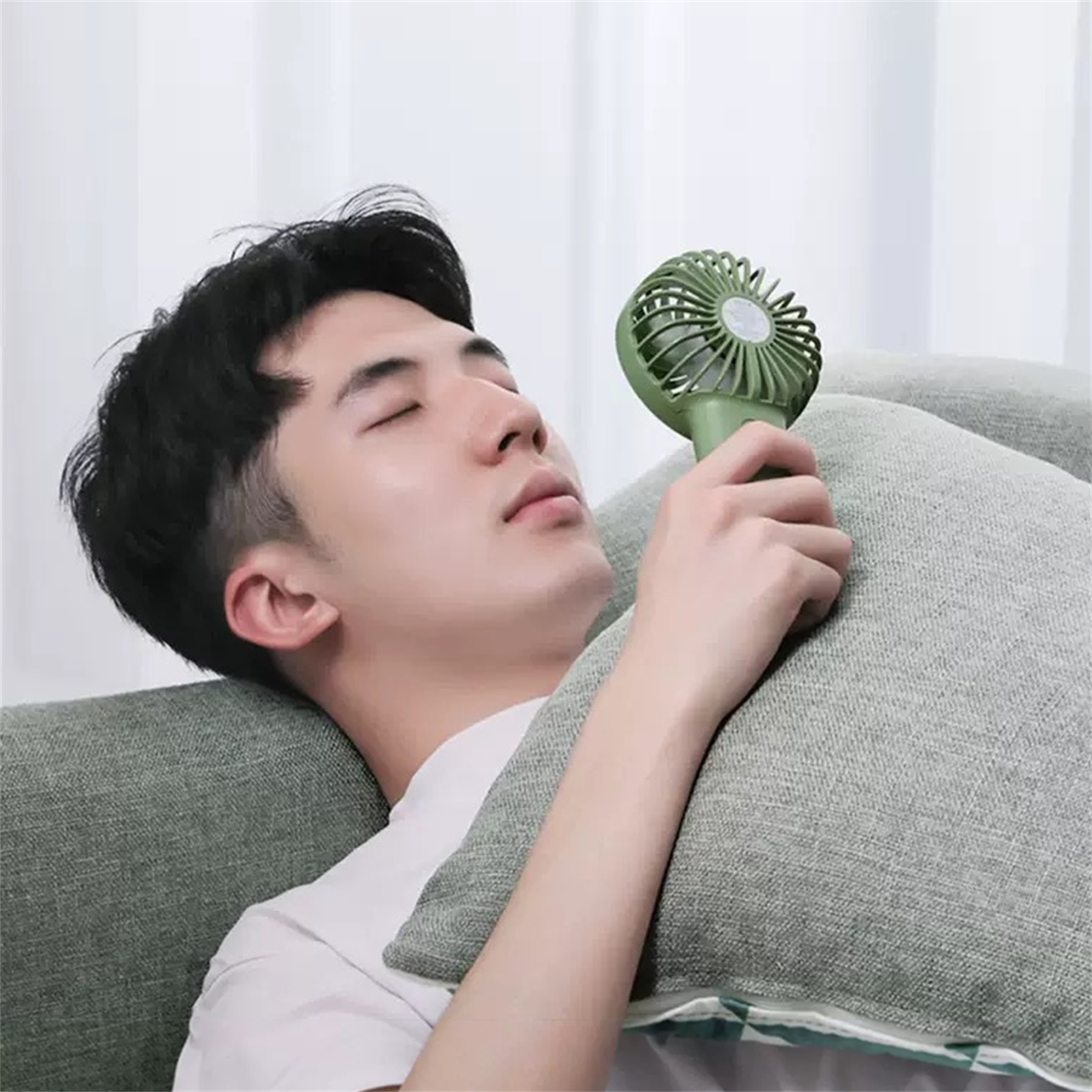 Ein Mann ruht auf einem Sofa mit einem Kissen und hält einen grünen Handventilator in der Nähe seines Gesichts.