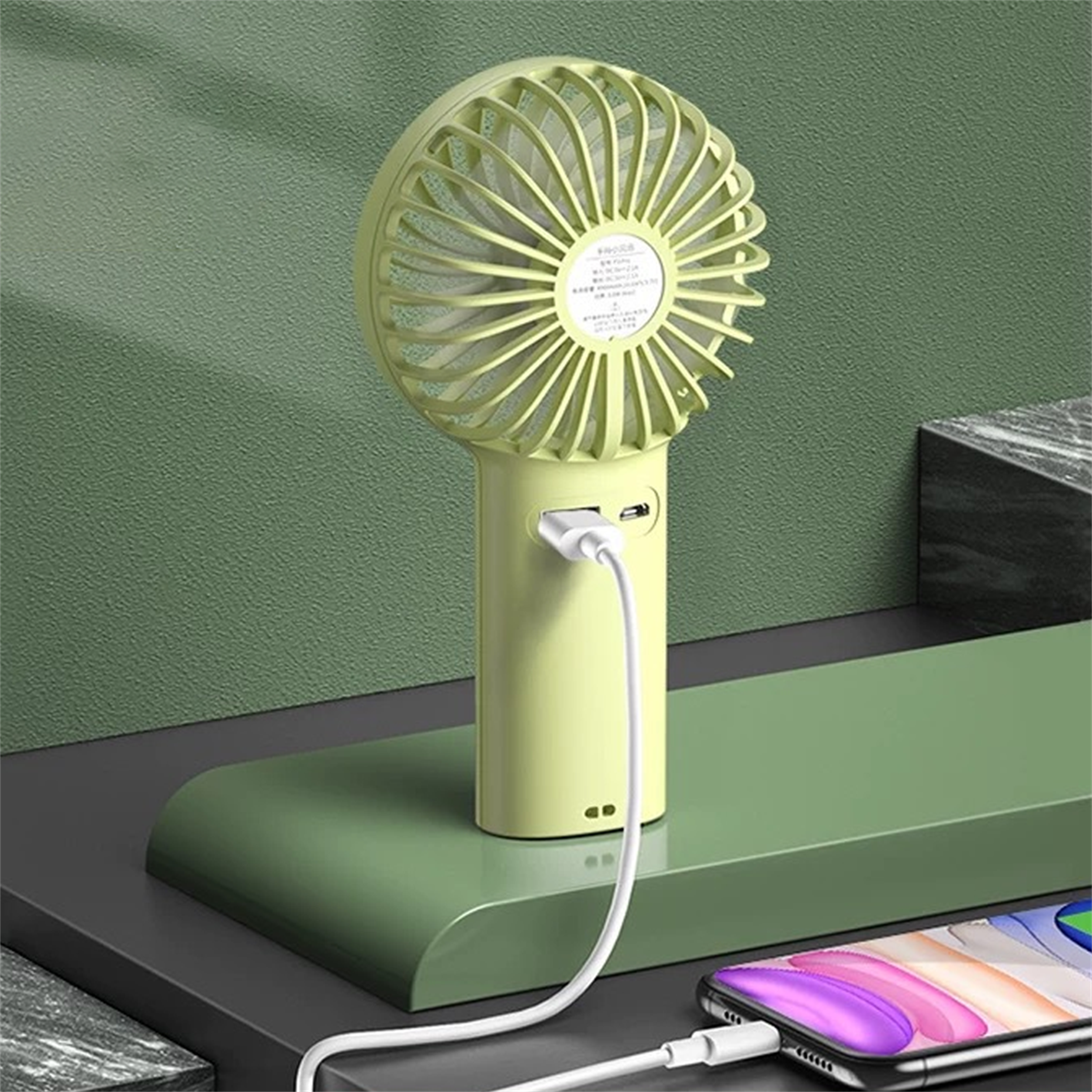 Handventilator, der mit einem Telefon geladen wird. Das Telefon wird ebenfalls geladen. Auf grüner Oberfläche.