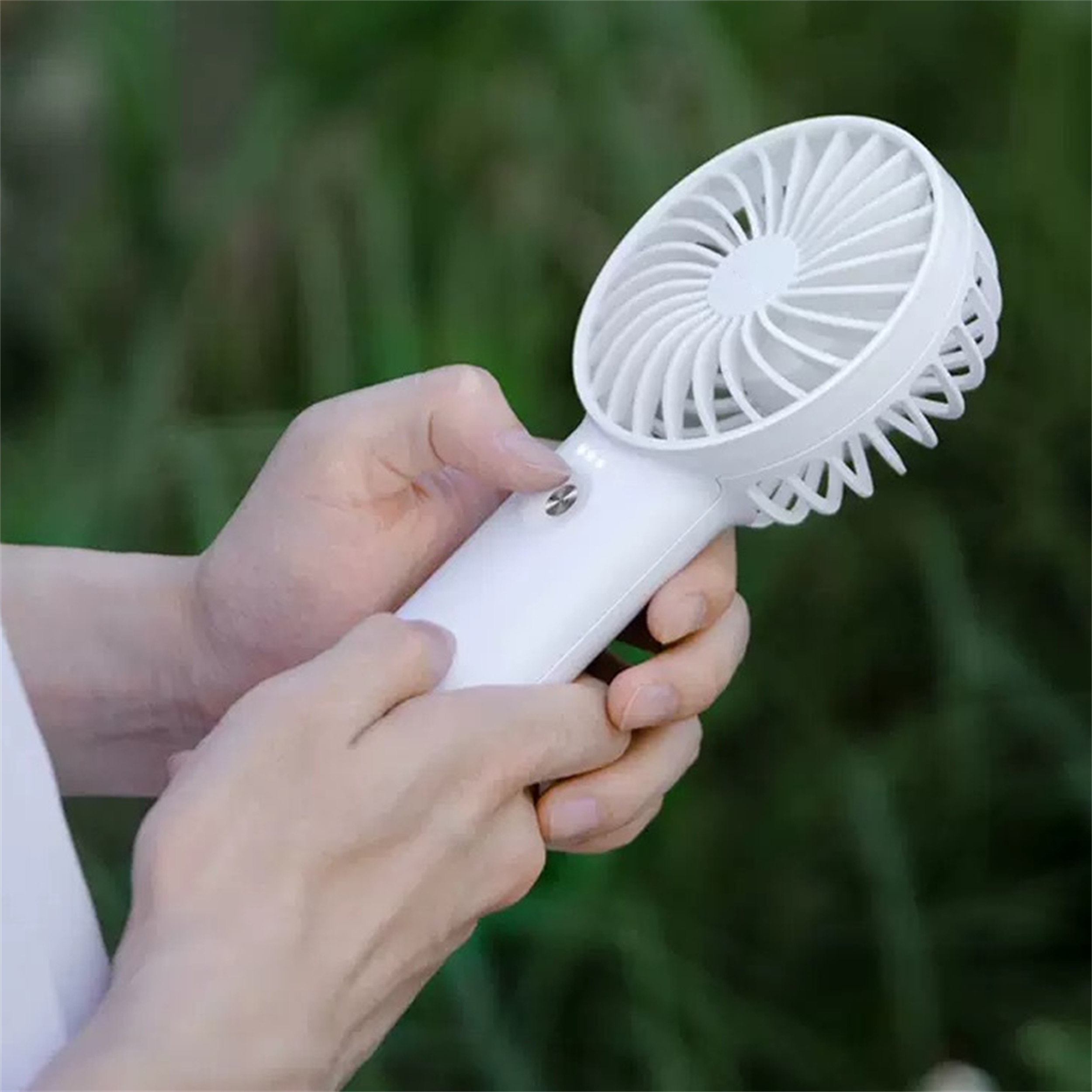 Hände halten einen weißen Handventilator vor verschwommenem grünen Hintergrund.