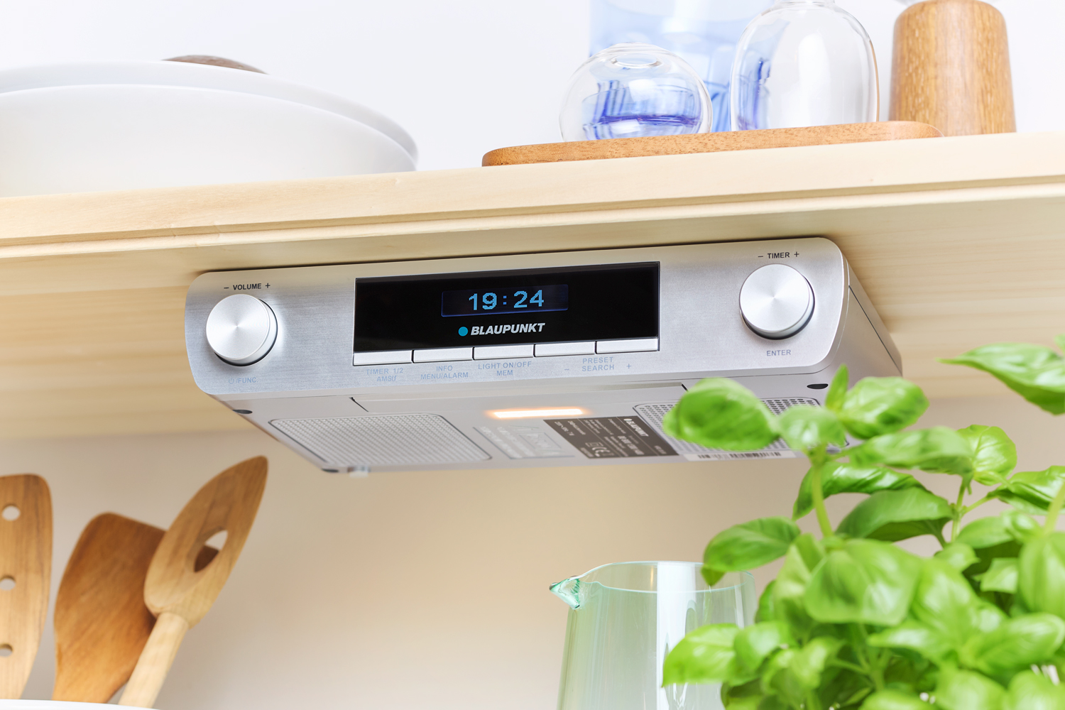 Ein silbernes Audiosystem unter einem Regal mit Uhrzeit (19:24) und Küchenumgebung im Hintergrund.