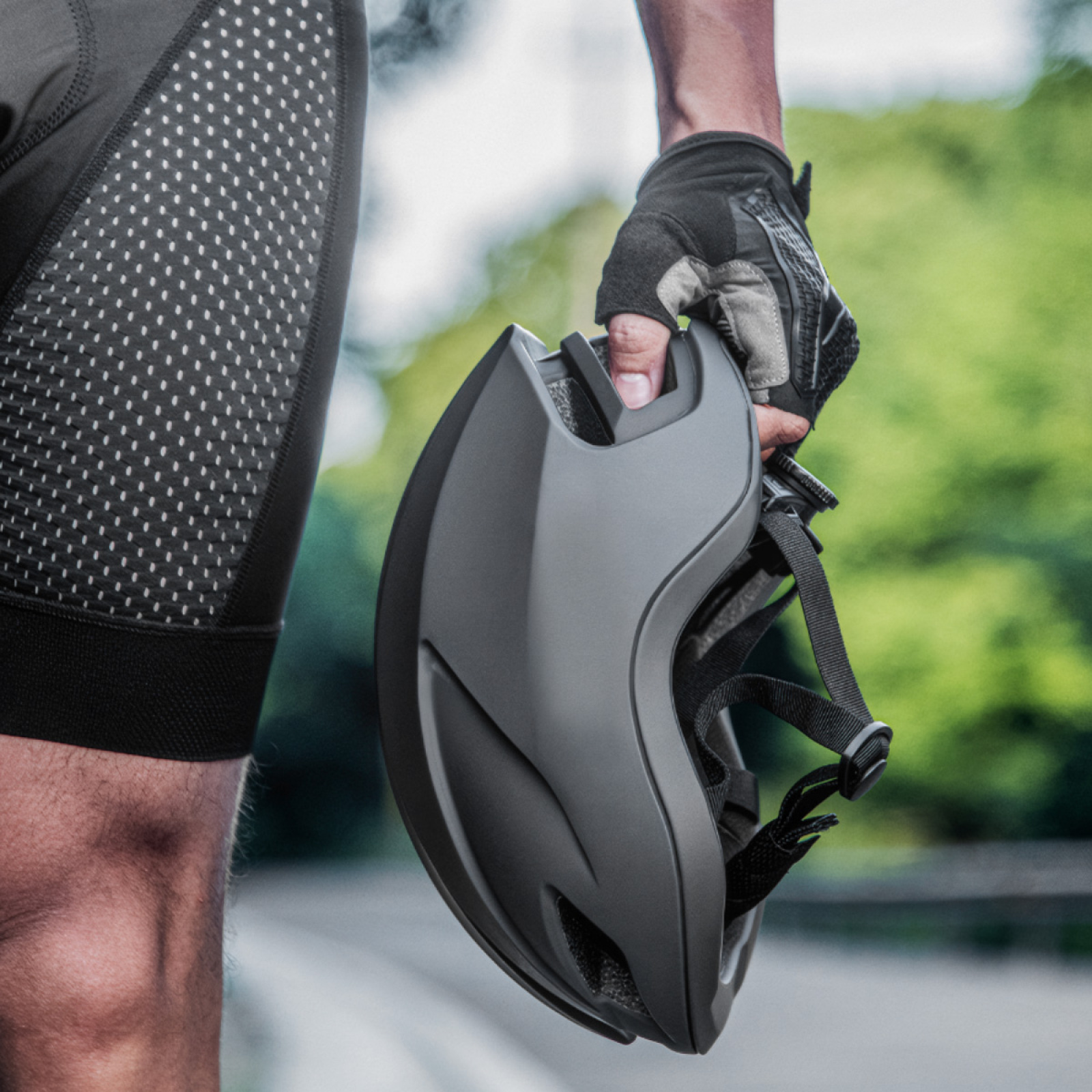 Hand hält einen dunkelgrauen Fahrradhelm. Person trägt Radlerhose.