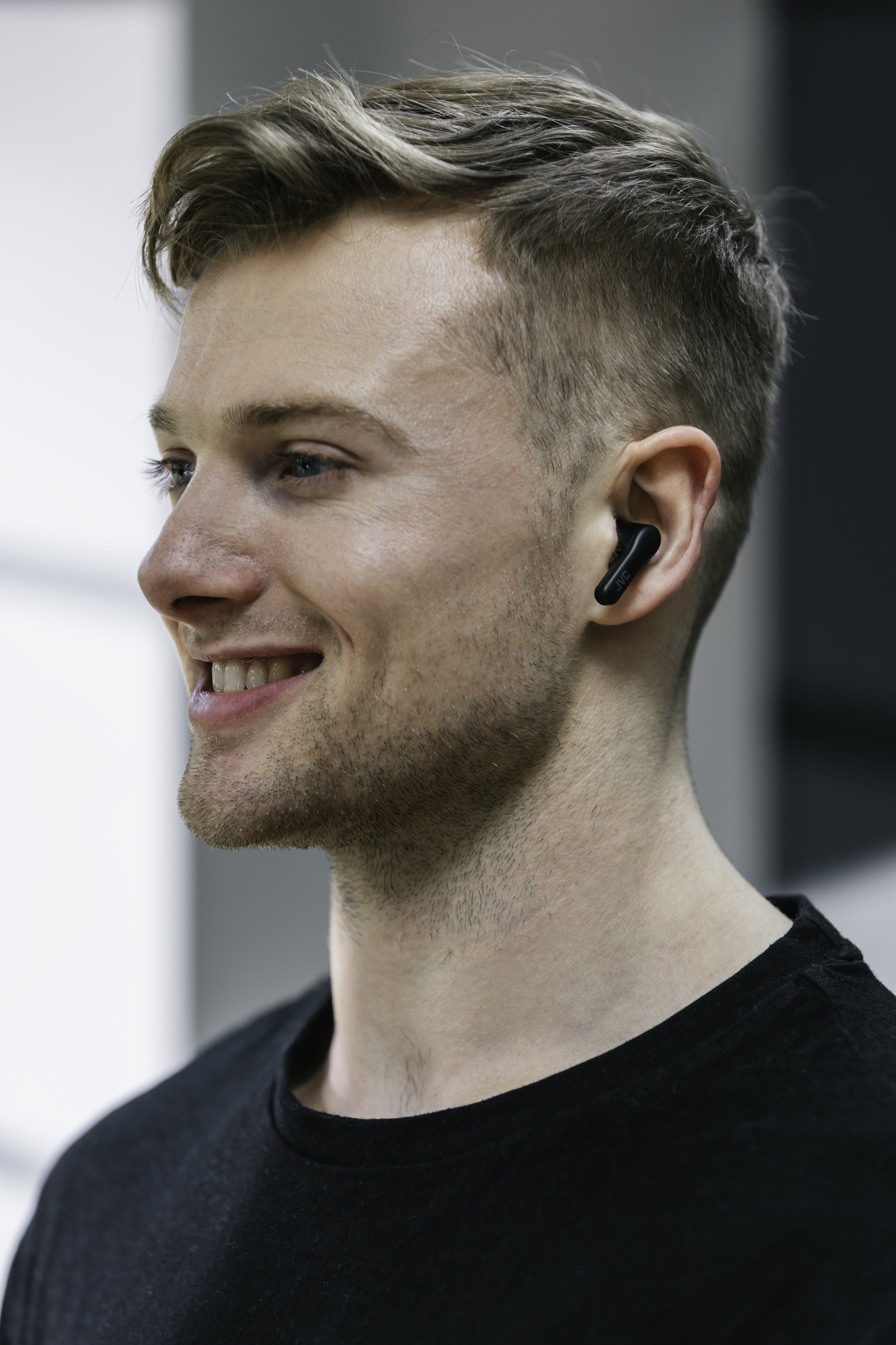 Mężczyzna uśmiecha się, nosząc czarne słuchawki douszne. Jest w profilu, patrzy w prawo.
