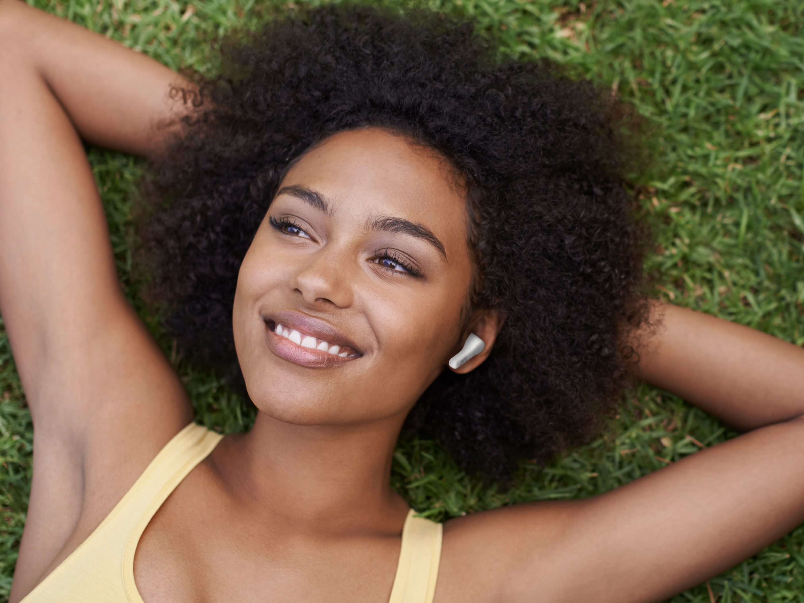 Kobieta leży na trawie w białych słuchawkach, patrząc w górę i uśmiechając się.