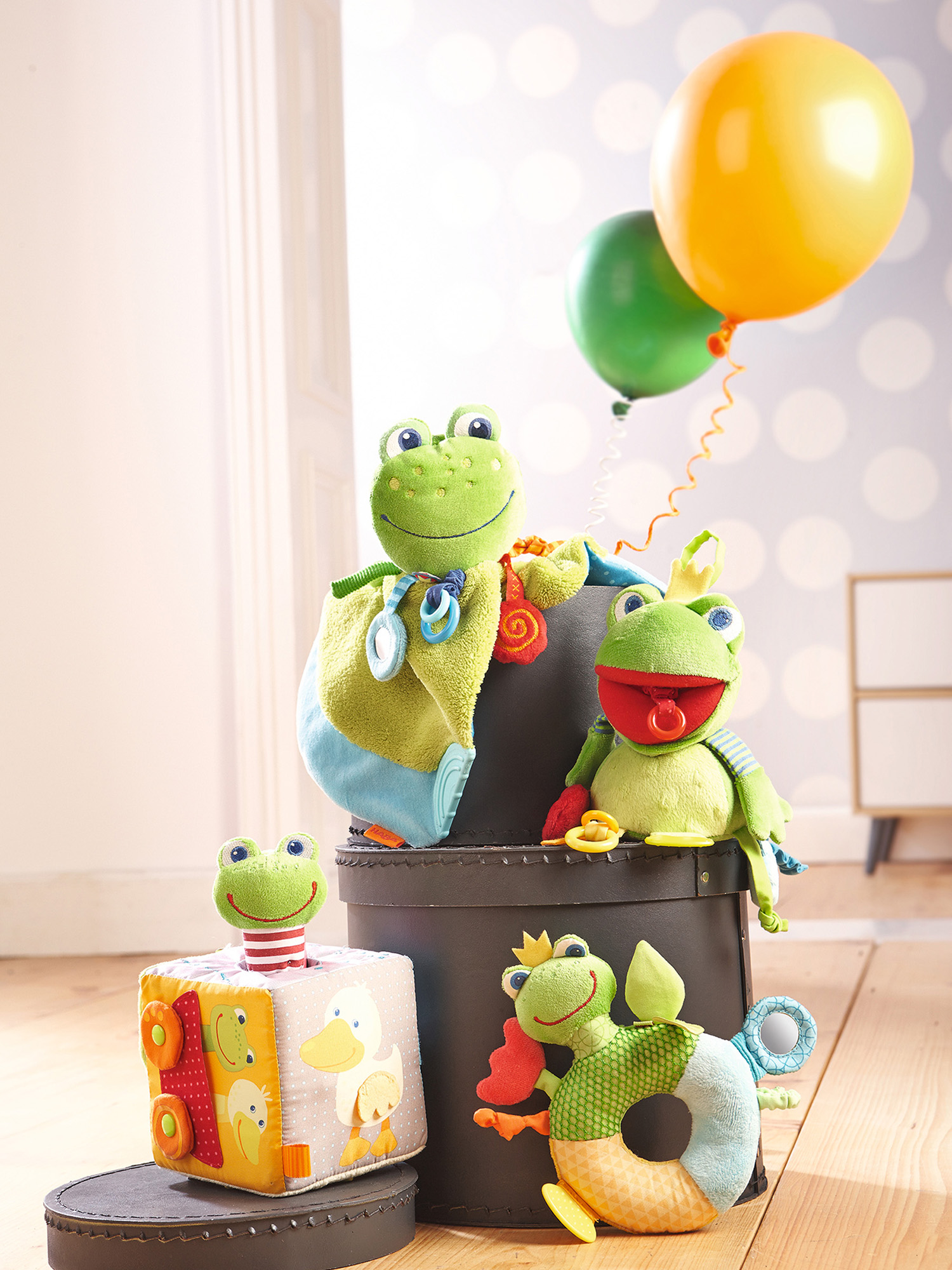 Grüne Froschspielzeuge auf einer schwarzen Box, mit Ballons im Hintergrund. Ein Spielzeugwürfel mit einer Ente darauf.