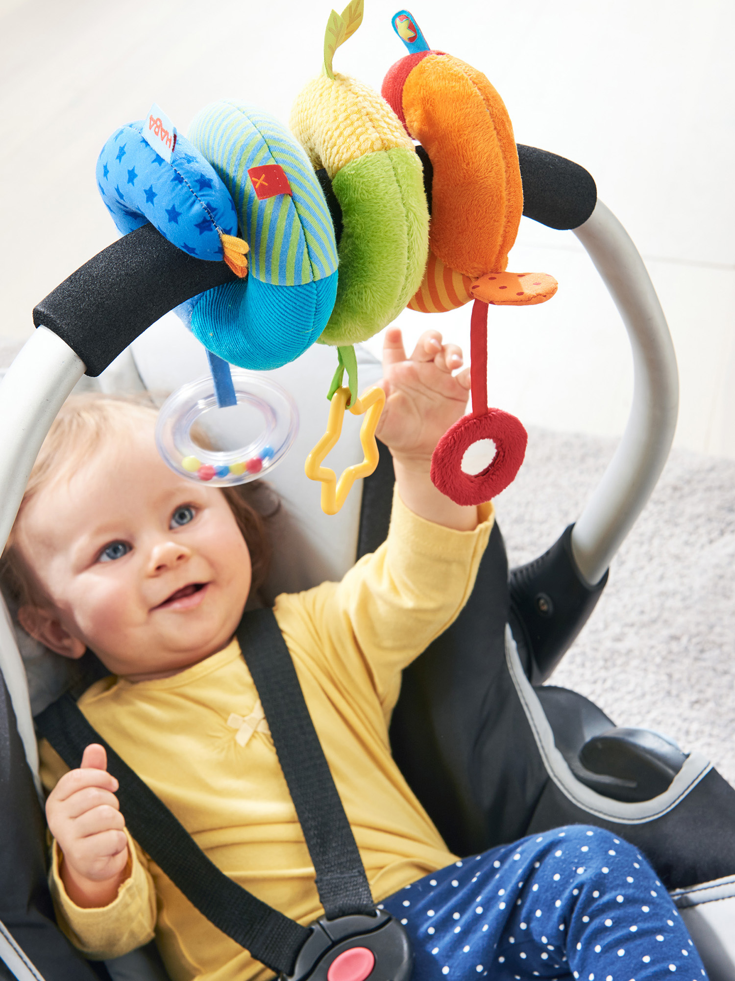 Ein Baby im Autositz greift nach einem bunten Spielzeug. Das Spielzeug hat verschiedene Formen und Farben.