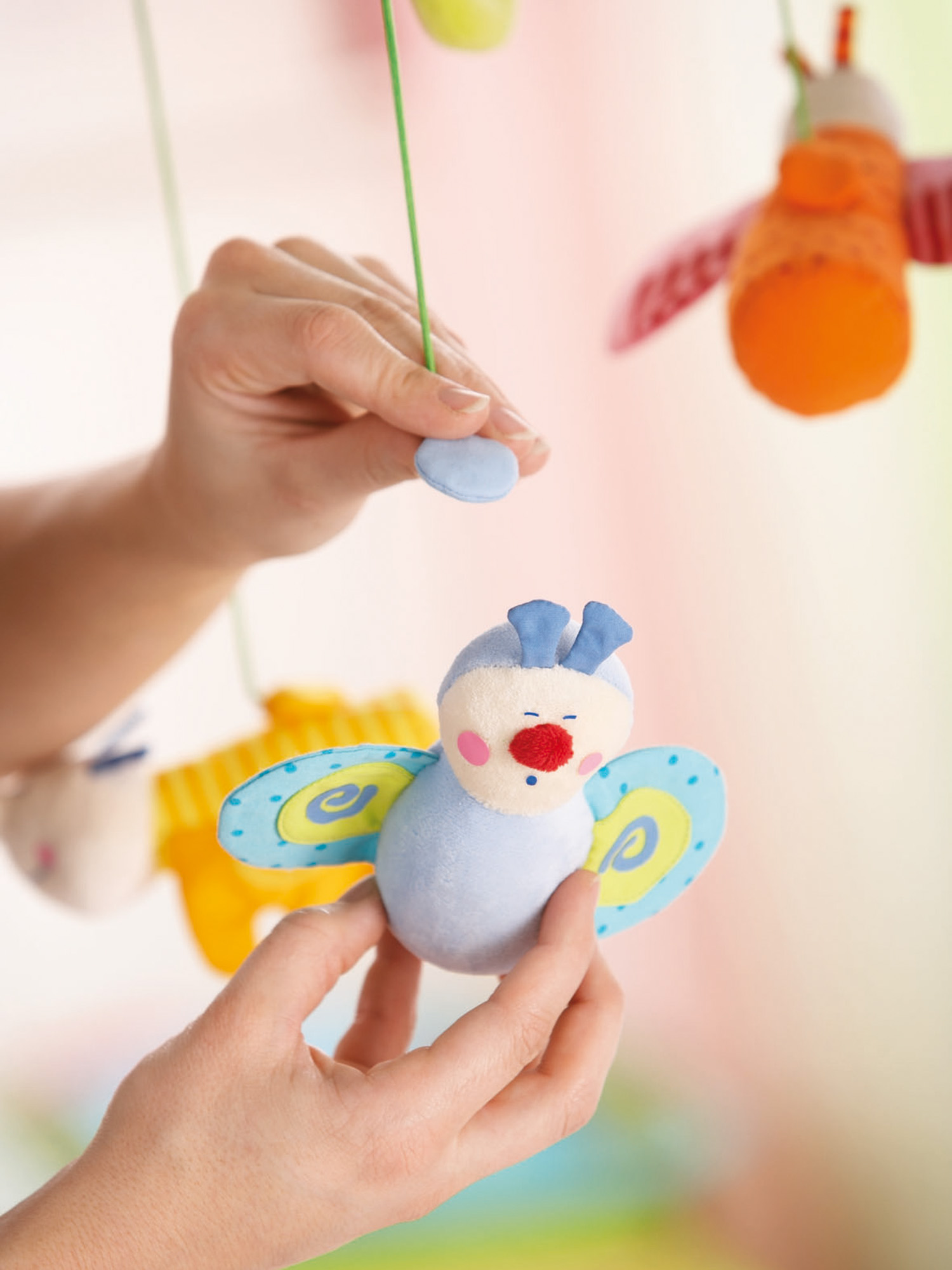 Hände halten ein Babymobile-Spielzeug mit bunten Stoffschmetterlingen und einer blaugesichtigen Biene.