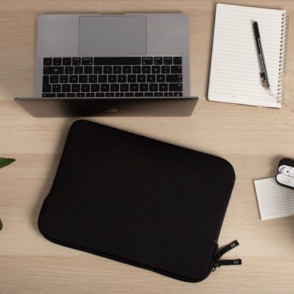 Une housse d'ordinateur portable noire sur une table en bois avec un ordinateur portable, un cahier et un stylo. Ouvert.