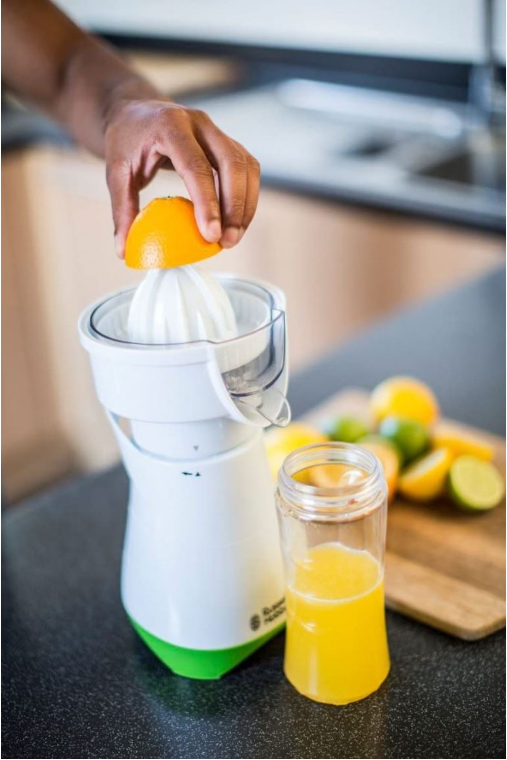 Person drückt Orangenhälfte in Entsafter. Saftglas und geschnittene Orangen auf dem Tisch. Weißer Entsafter.