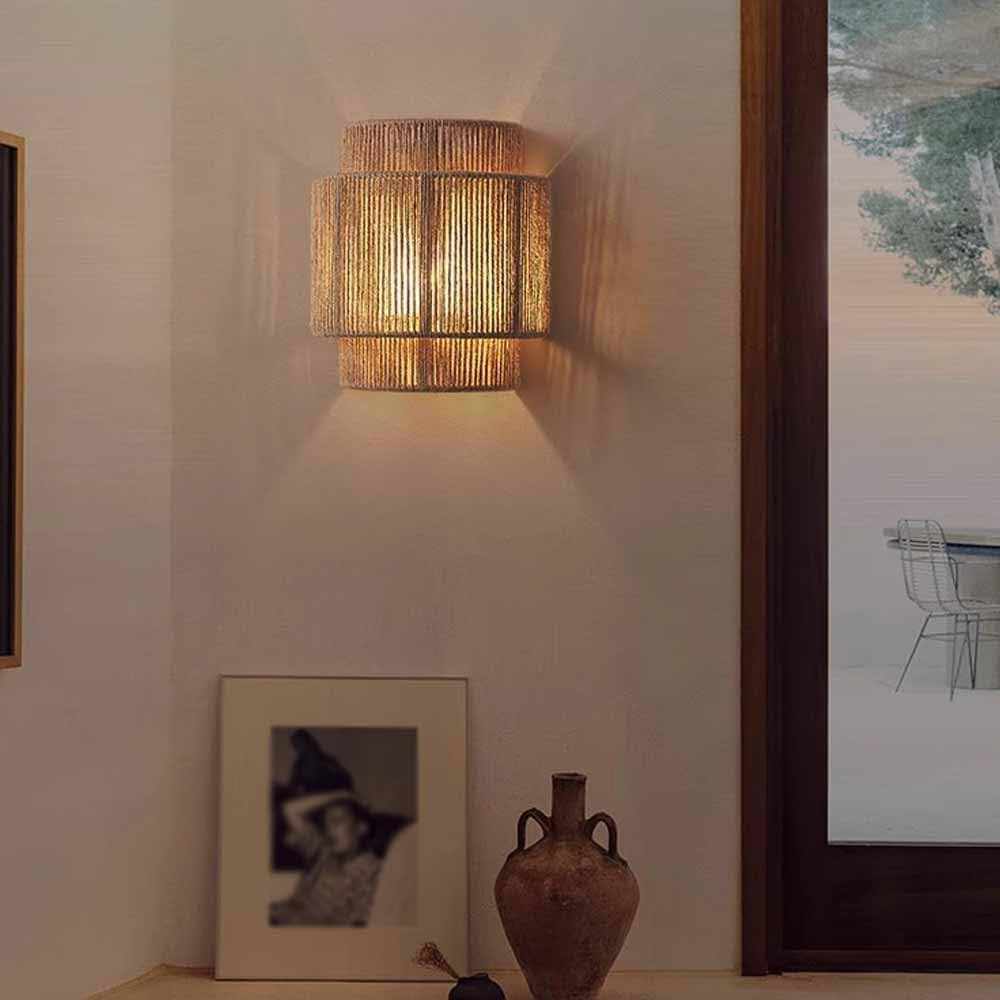 Lámpara de pared con pantalla tejida, emitiendo luz cálida. Un cuadro y un jarrón en primer plano.
