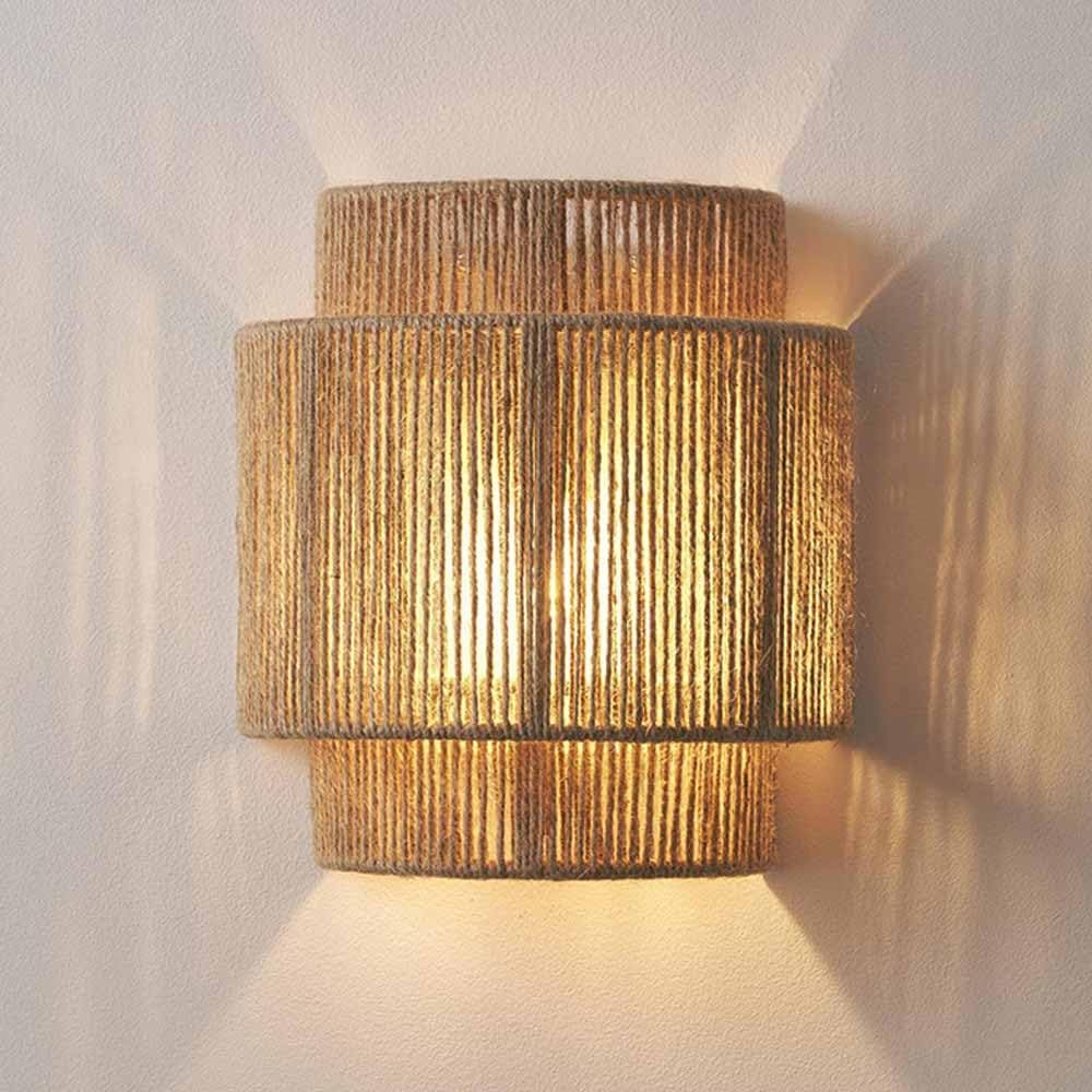 Lámpara de pared con pantalla cilíndrica tejida, emitiendo luz cálida. La pared es blanquecina.