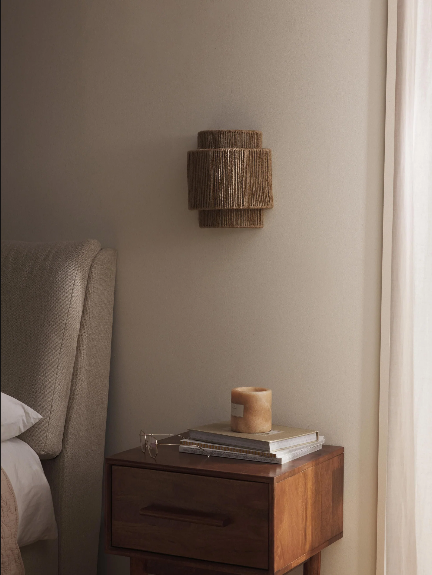 Una pared con una lámpara de pared, una mesita de noche con libros y una vela, y una cama.