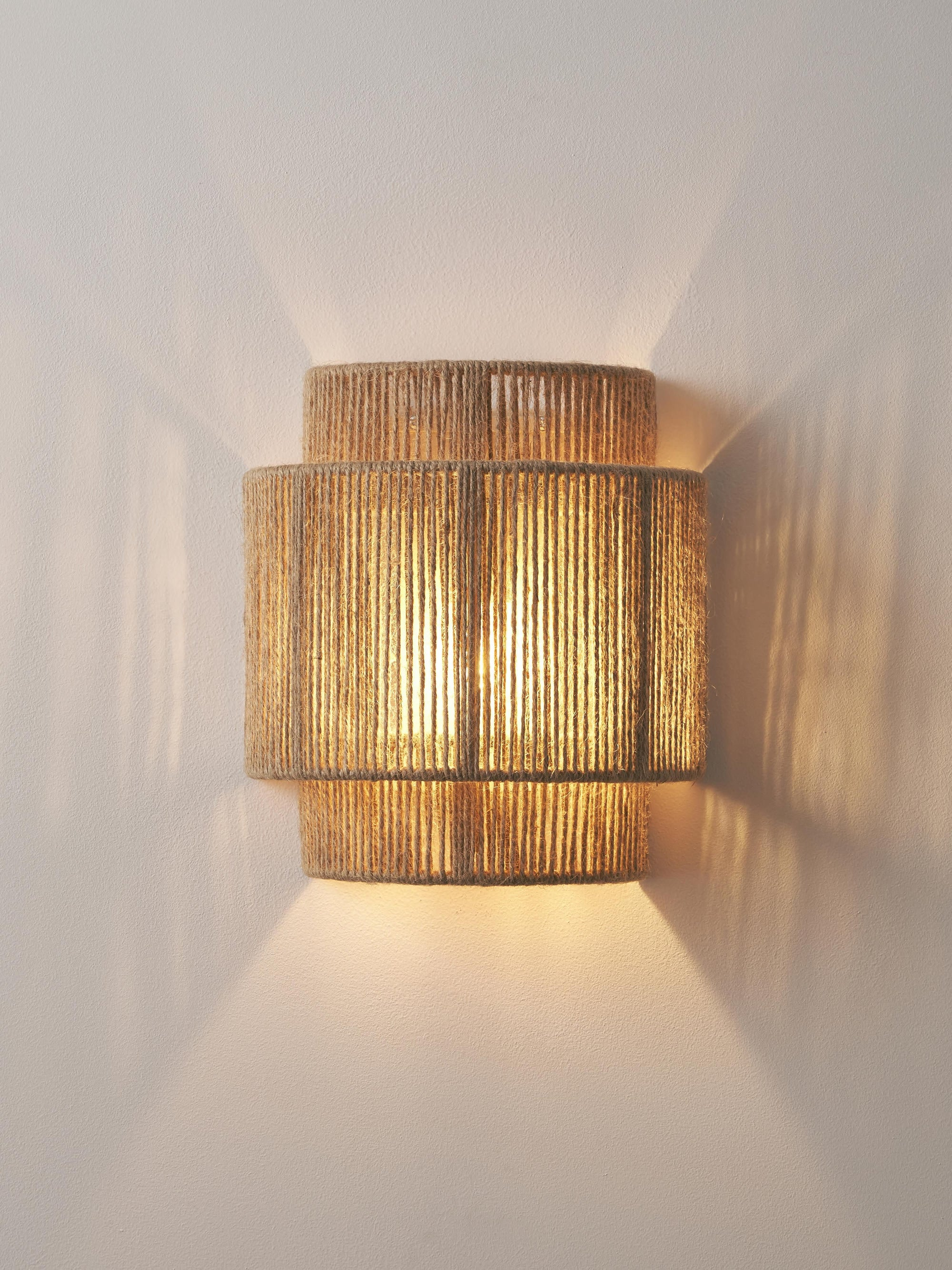 Una lámpara de pared con un diseño tejido en capas, que proyecta un brillo cálido en la pared. La luz es suave y enfocada.
