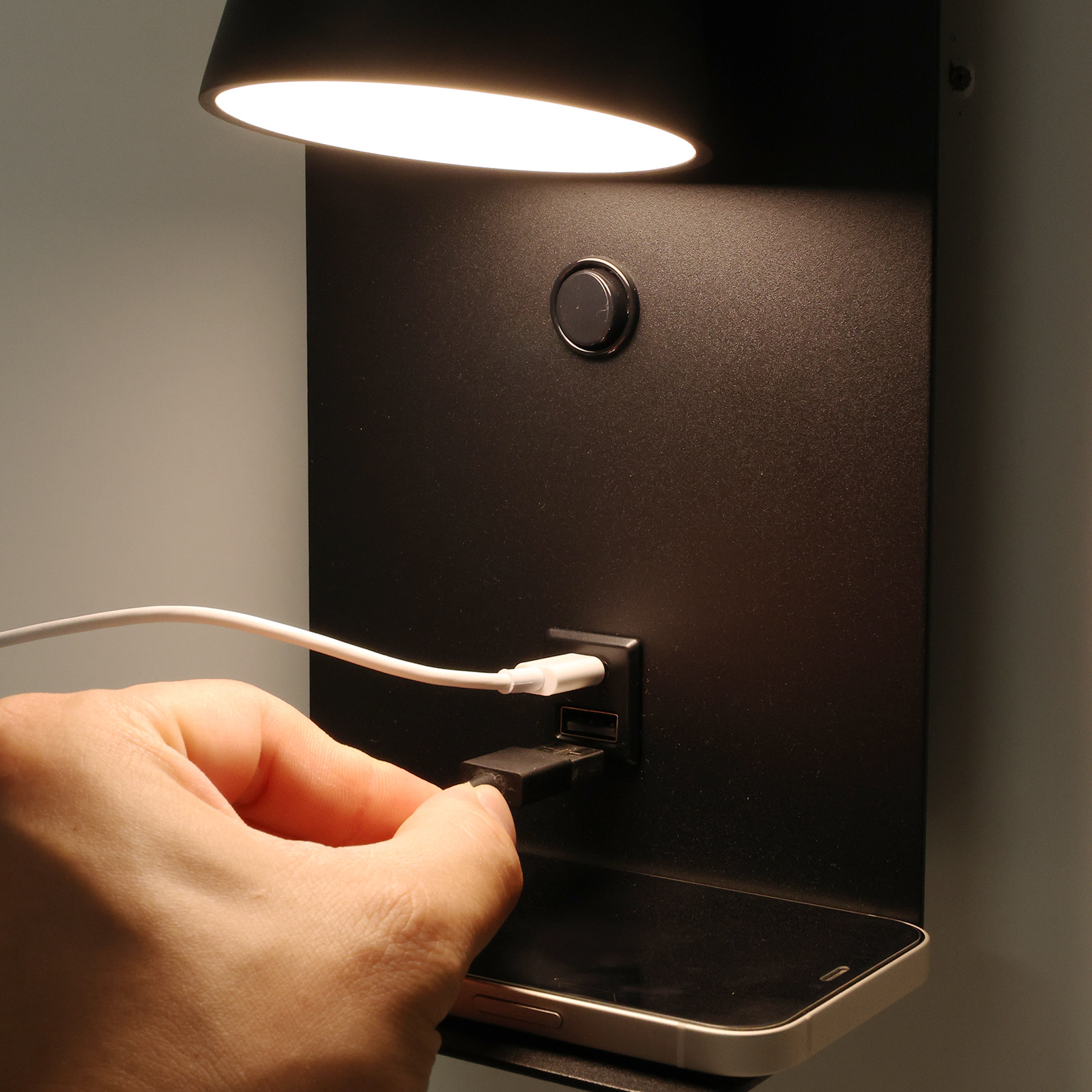 Una lámpara de pared negra con puerto de carga USB. Se ve una mano insertando un cable USB. La lámpara tiene un estante.