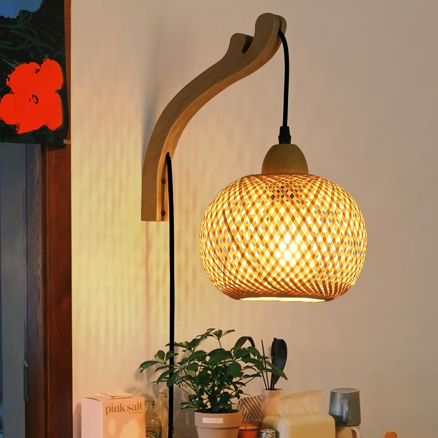 Una lámpara de pared de madera con una pantalla esférica tejida y un cable negro. Cuelga junto a una pared con una flor roja.