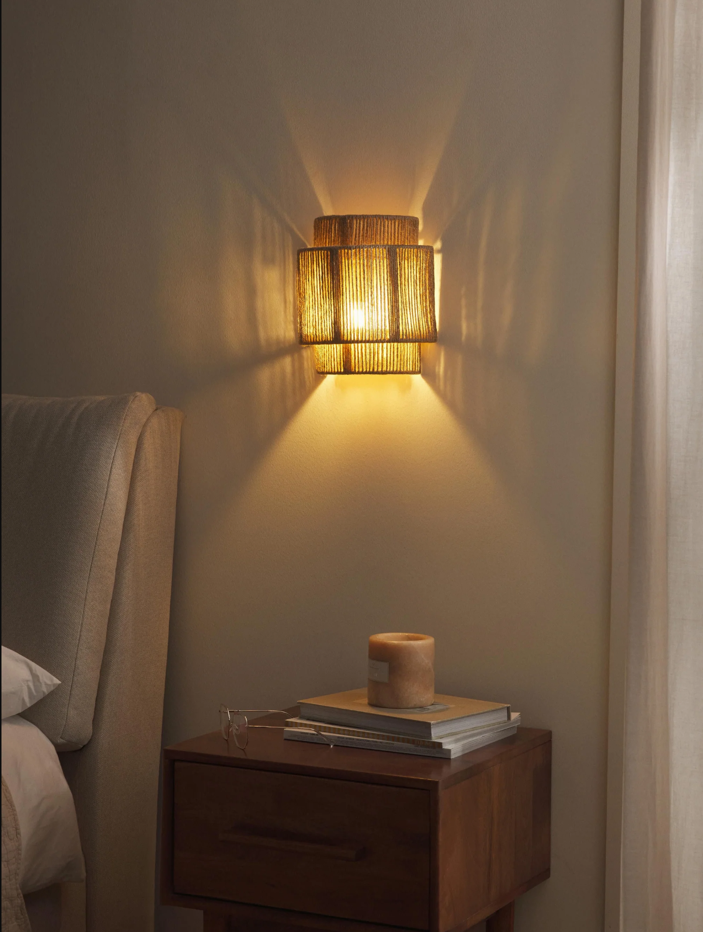 Una lámpara de pared con una pantalla tejida está montada en una pared beige sobre una mesita de noche.