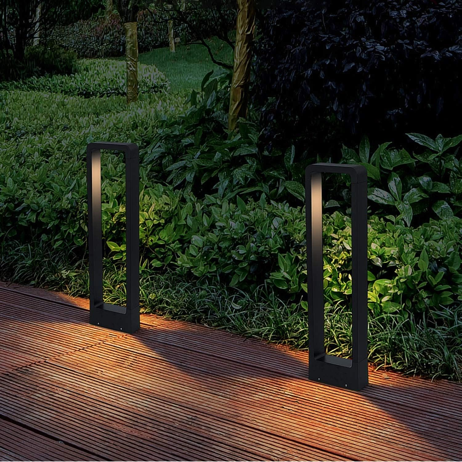 Dos lámparas rectangulares negras en una terraza de madera, emitiendo luz cálida, con arbustos verdes detrás.