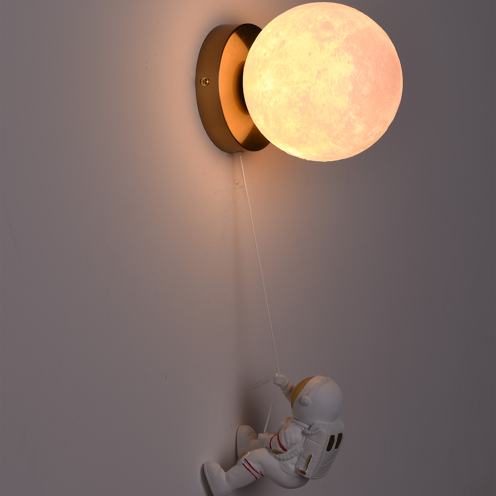 Lámpara de pared con forma de luna y figura de astronauta colgando. Fondo gris claro.