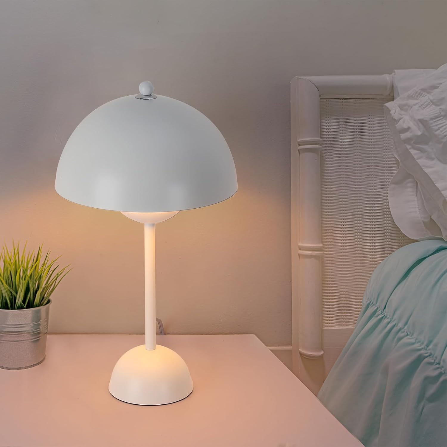 Una lámpara blanca con una pantalla en forma de hongo está sobre una mesa blanca. Una planta a la izquierda.