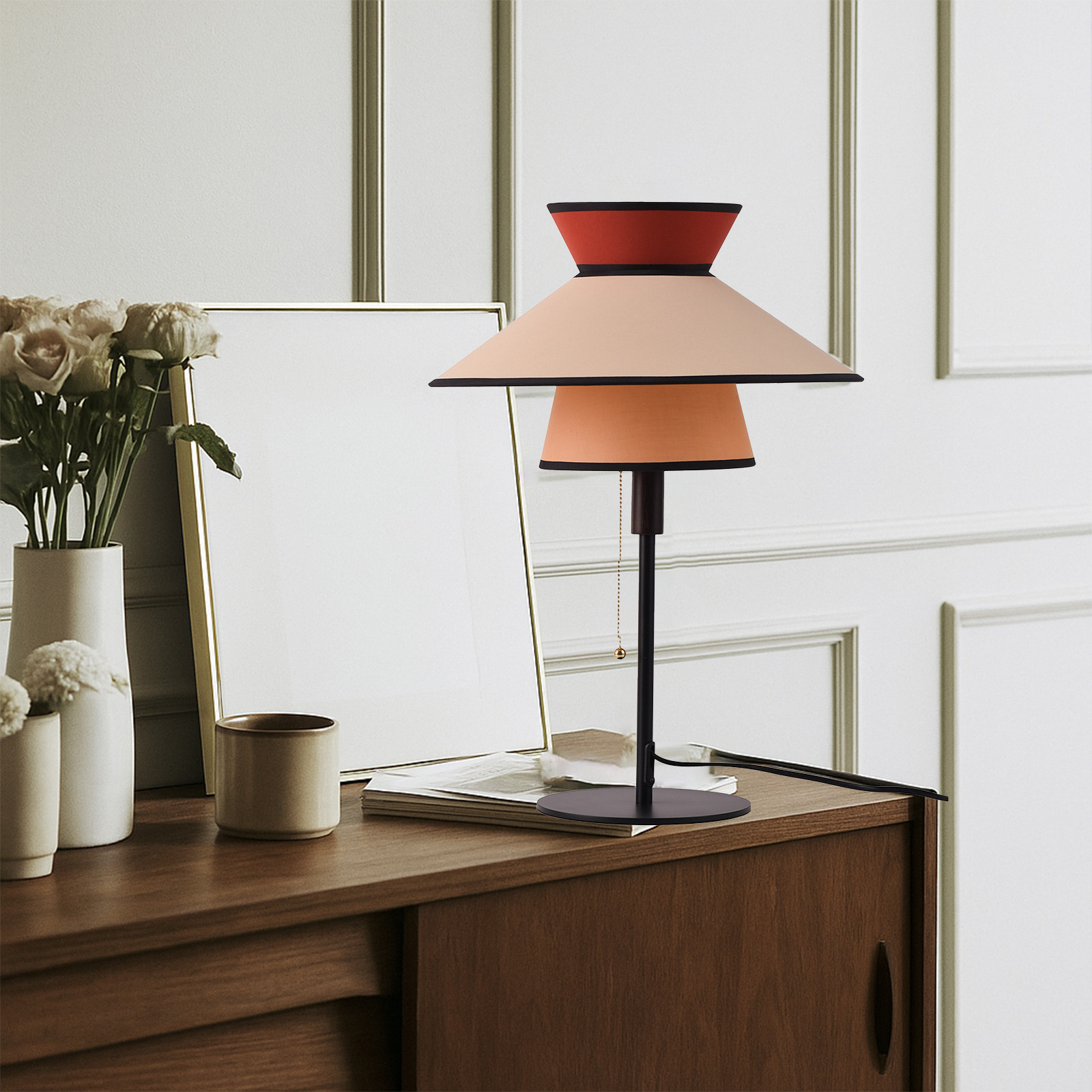 Una lámpara de mesa con base negra, dos pantallas y pantalla superior roja, junto a un jarrón de rosas.