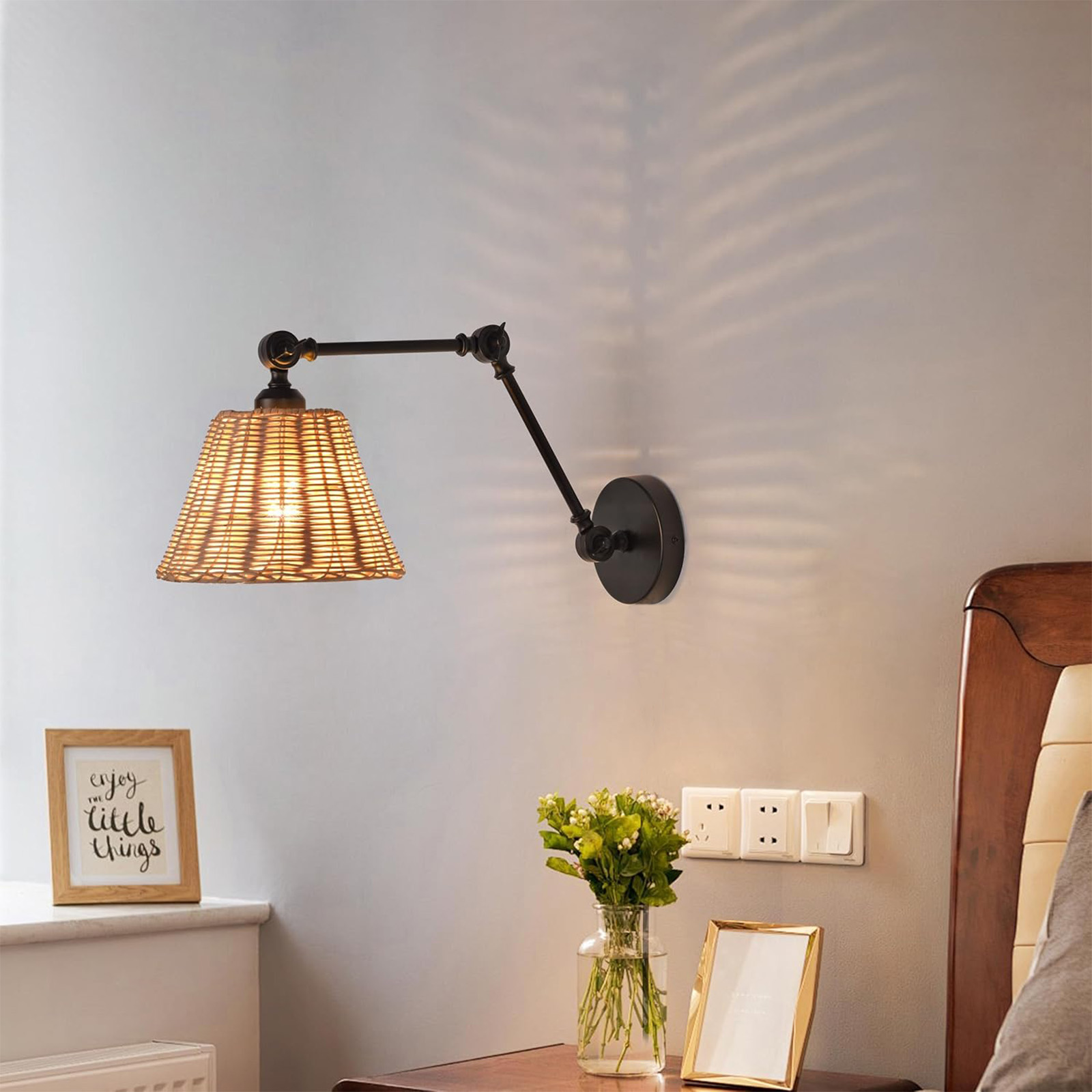 Una lámpara de pared con una pantalla tejida, iluminada, junto a una cama, un jarrón con flores, un marco.