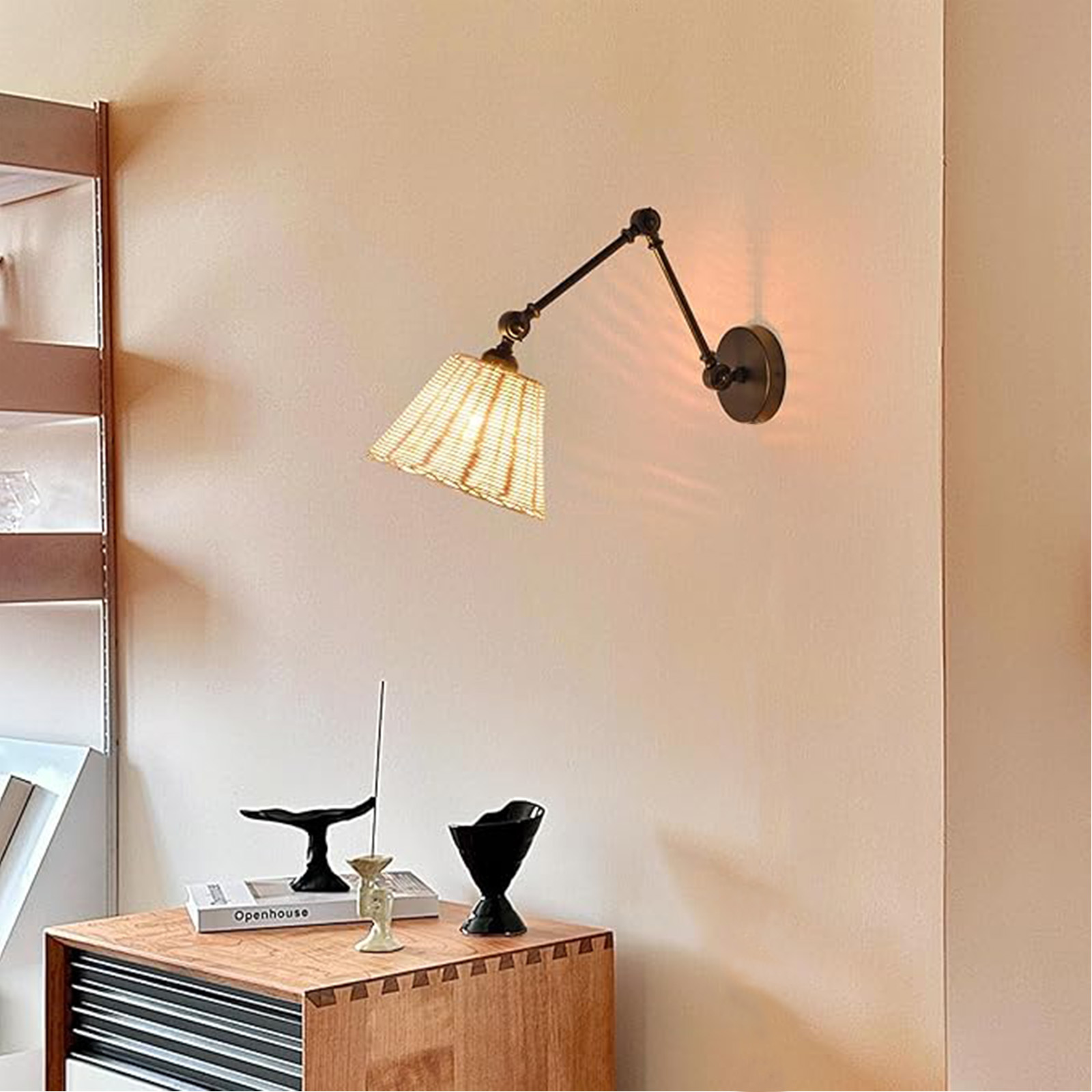 Una lámpara de pared con una pantalla tejida, encendida, proyectando luz sobre una mesa con elementos decorativos.
